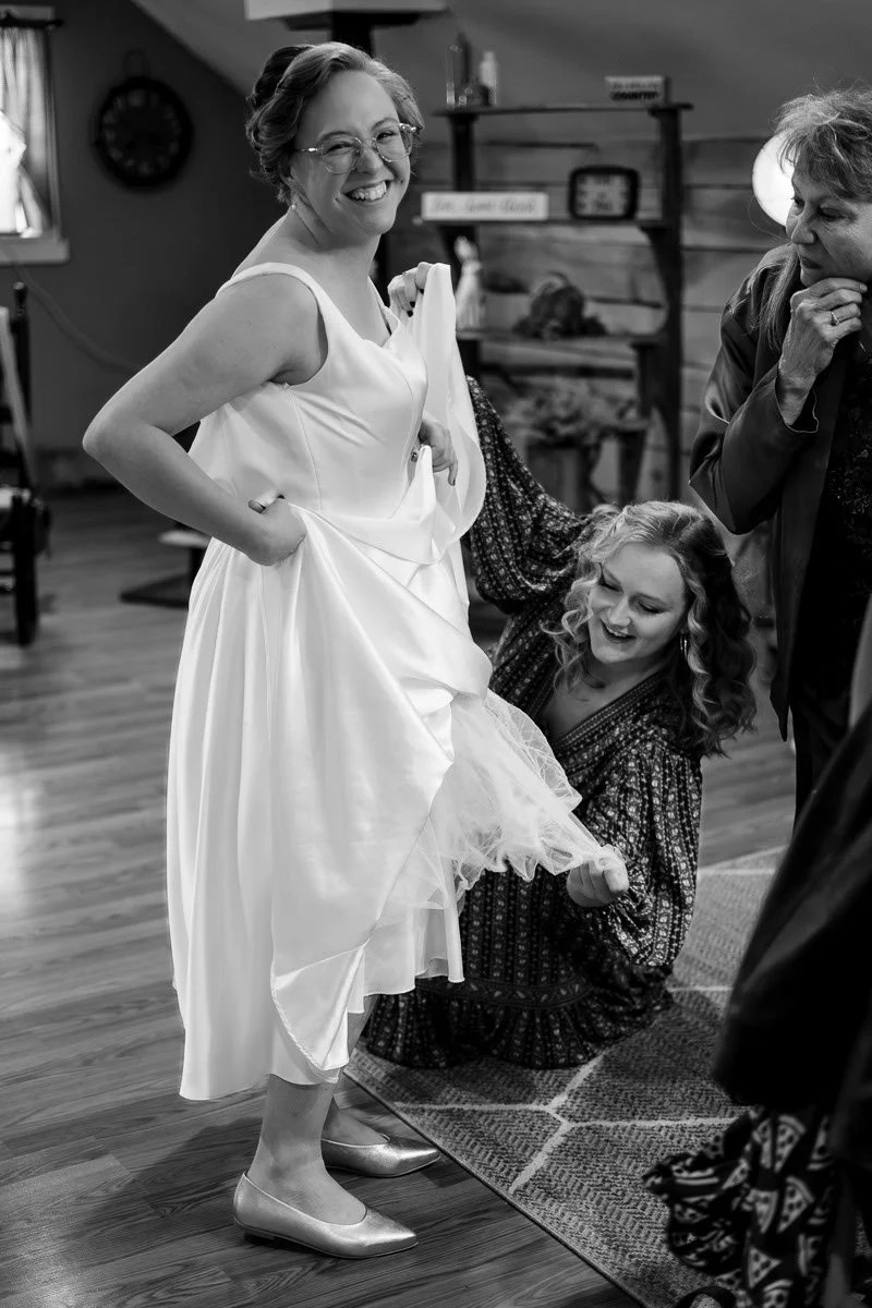 Woman in a wedding dress smiling, surrounded by friends in a cozy, rustic indoors setting.