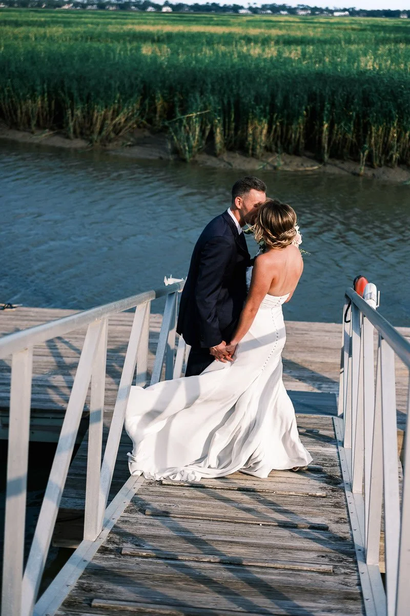 Bride and groom holding hands on a dock by a river, with a green field and trees in the background.