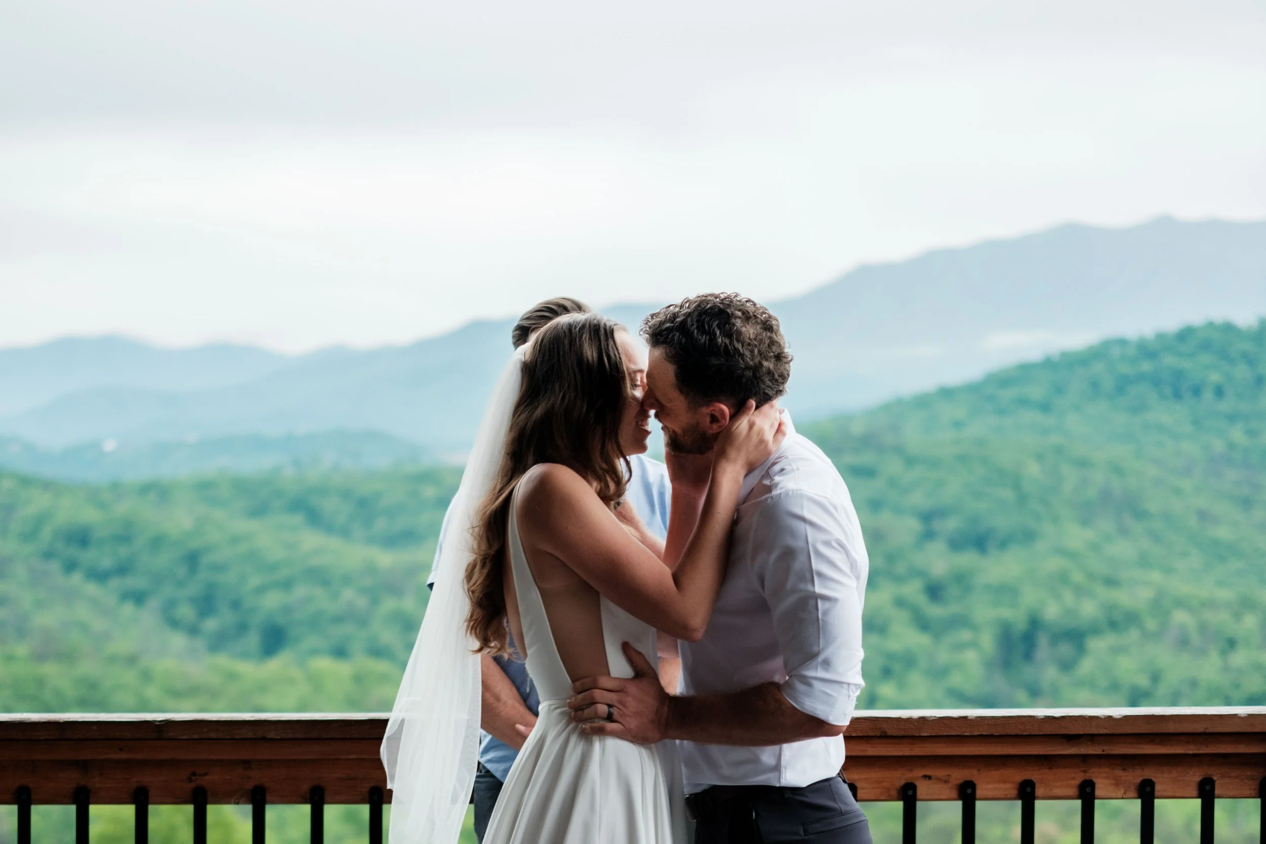 A couple sharing a loving embrace and kiss on a balcony overlooking a lush green mountain landscape during daytime.