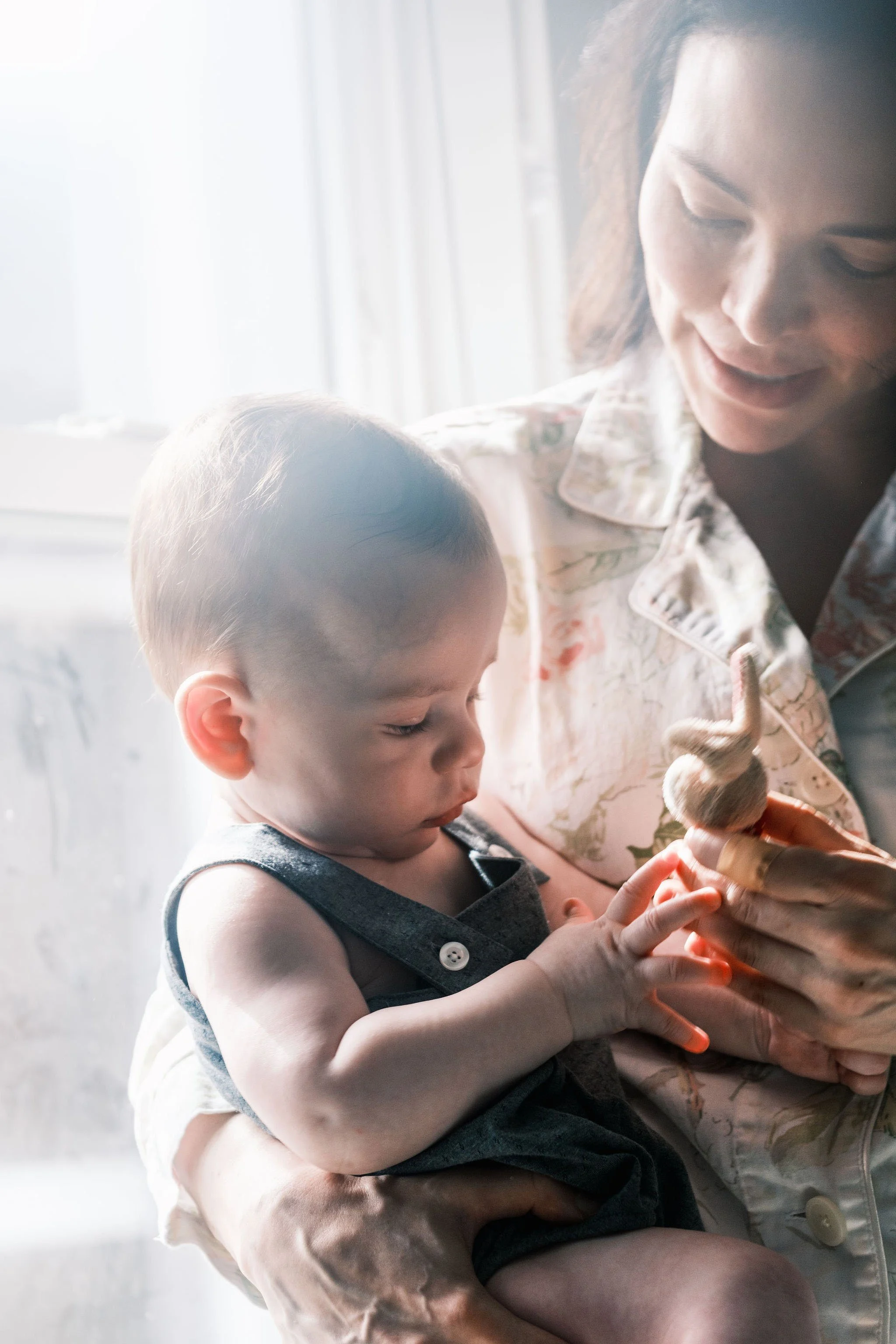 A woman holding a young boy who is touching a small lizard on her hand, near a bright window.