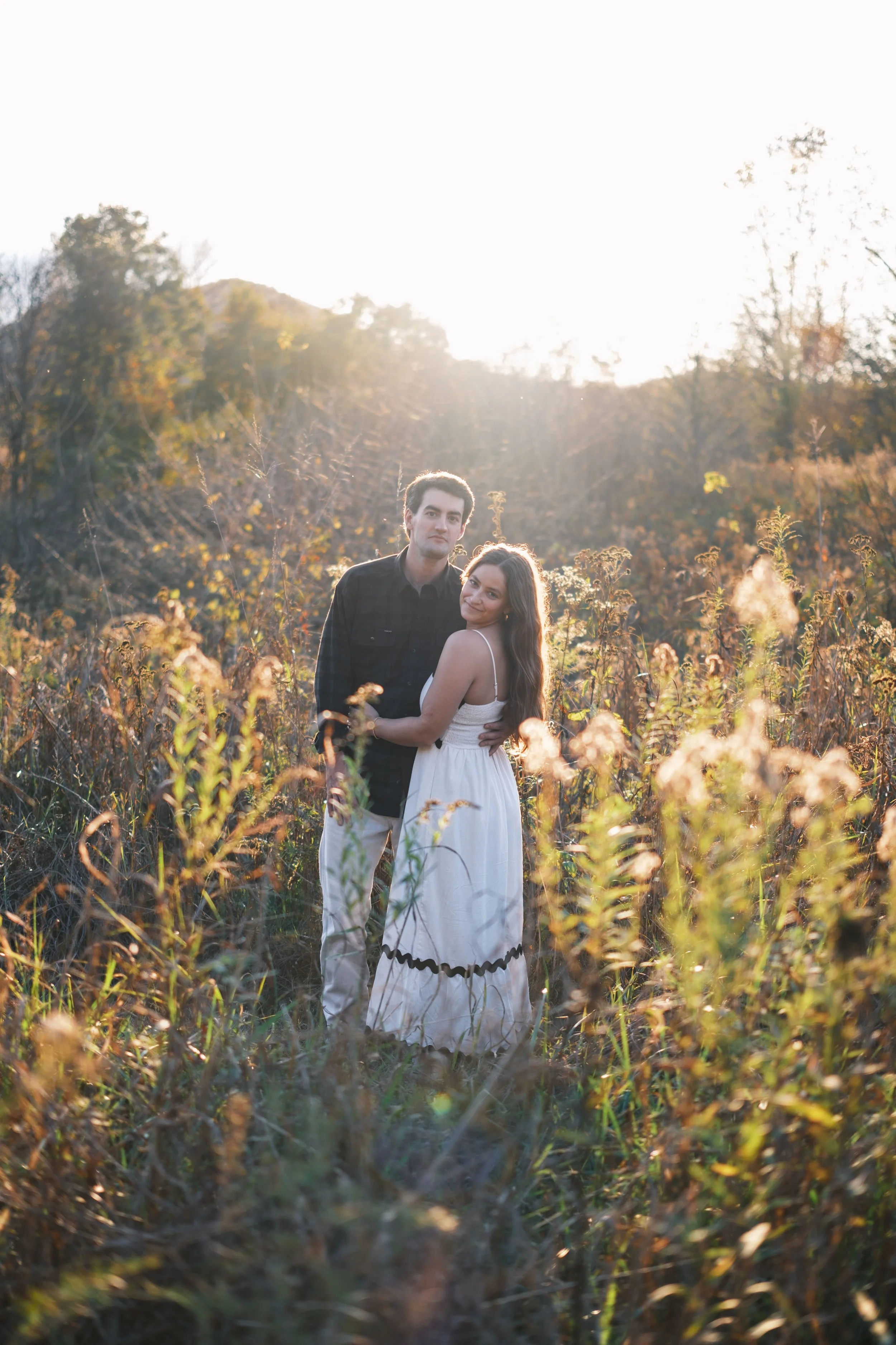 A couple standing in a field of tall grass and wildflowers during sunset, embracing each other and looking at the camera.
