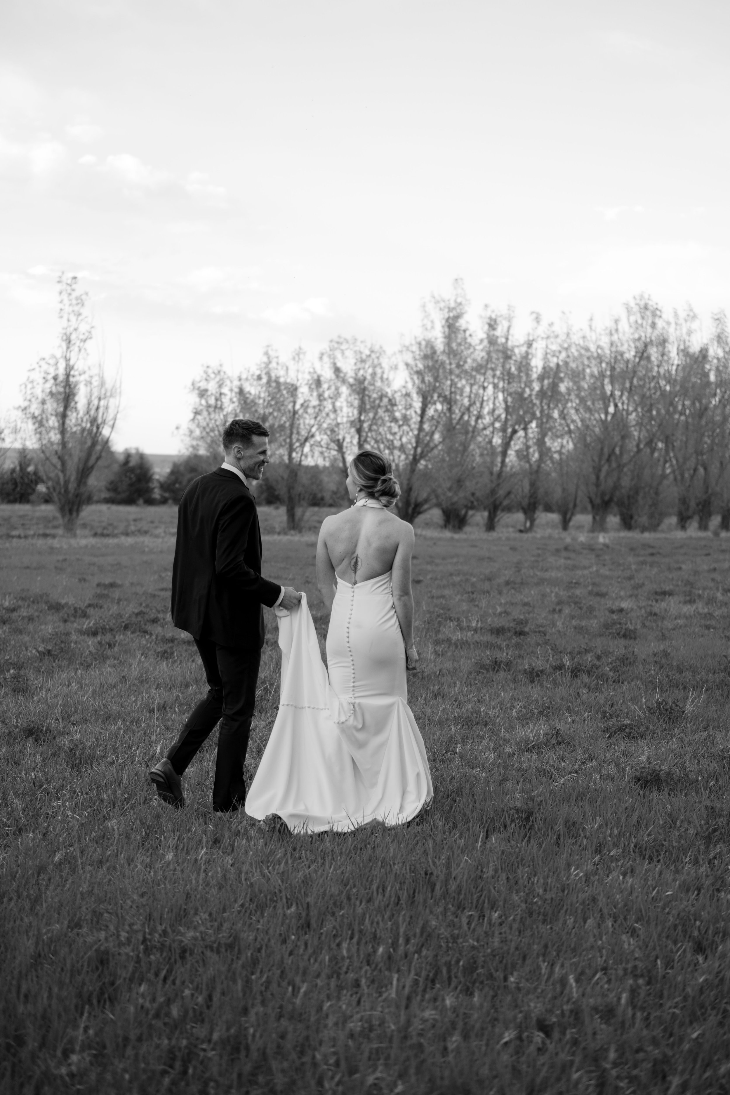 Black and white photo of a bride and groom walking in a grassy field, holding the bride's train, with trees in the background.