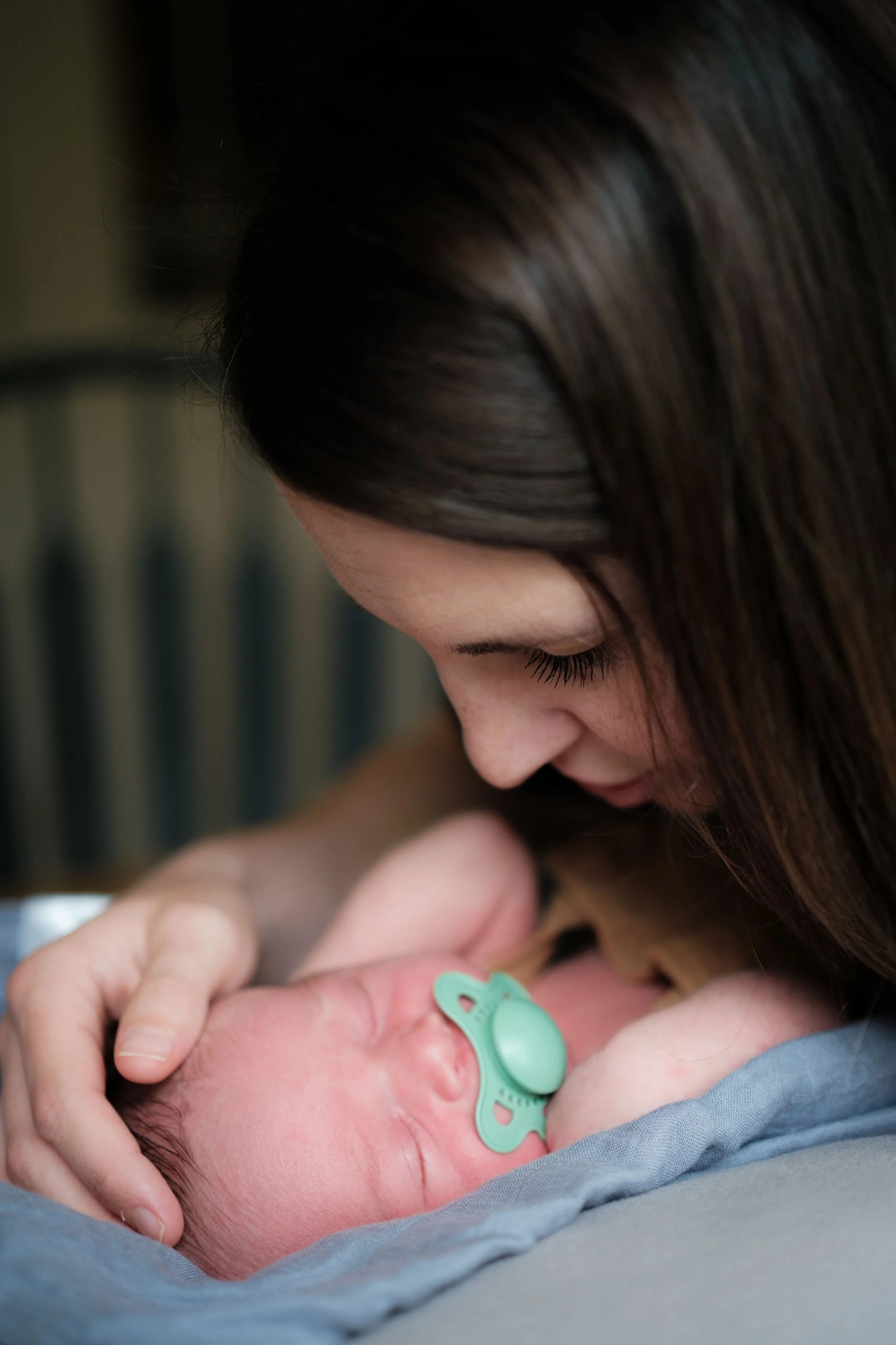 Woman gently holding and leaning over a sleeping newborn baby with a pacifier, lying on a blanket.