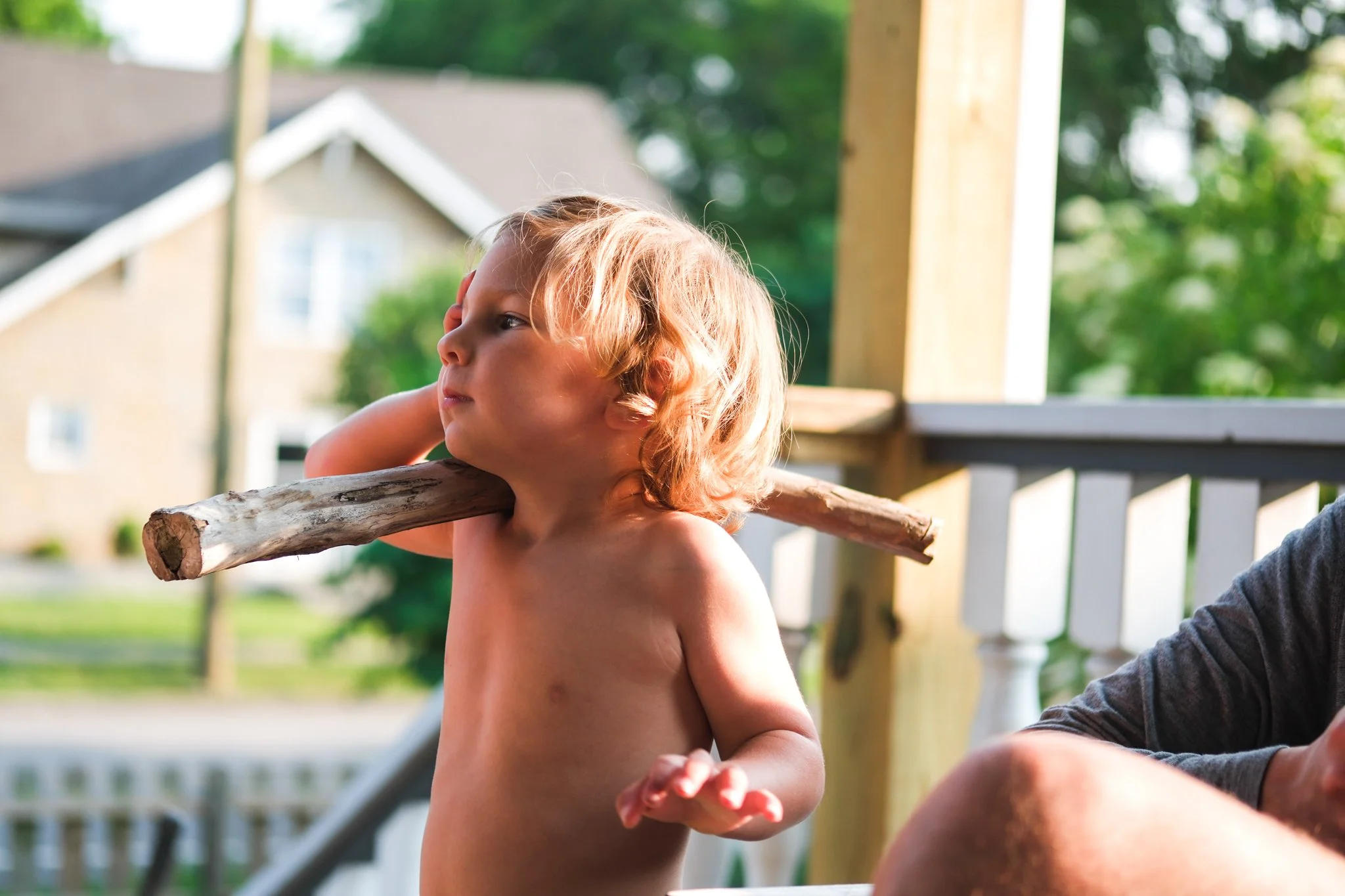 A young, shirtless boy with blonde, wavy hair holds a large stick on his shoulder and looks to the side. He is outdoors on a porch or deck, with a house and green trees in the background.
