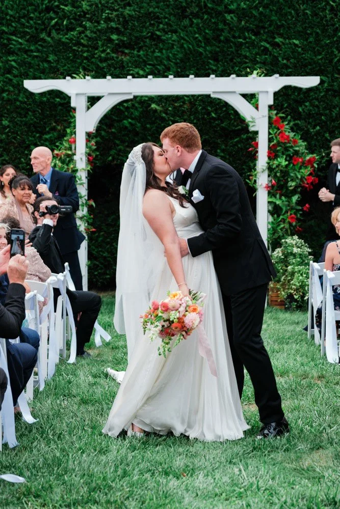 Bride and groom kissing during their outdoor wedding ceremony, surrounded by guests sitting on white chairs, with a green hedge and floral arch in the background.