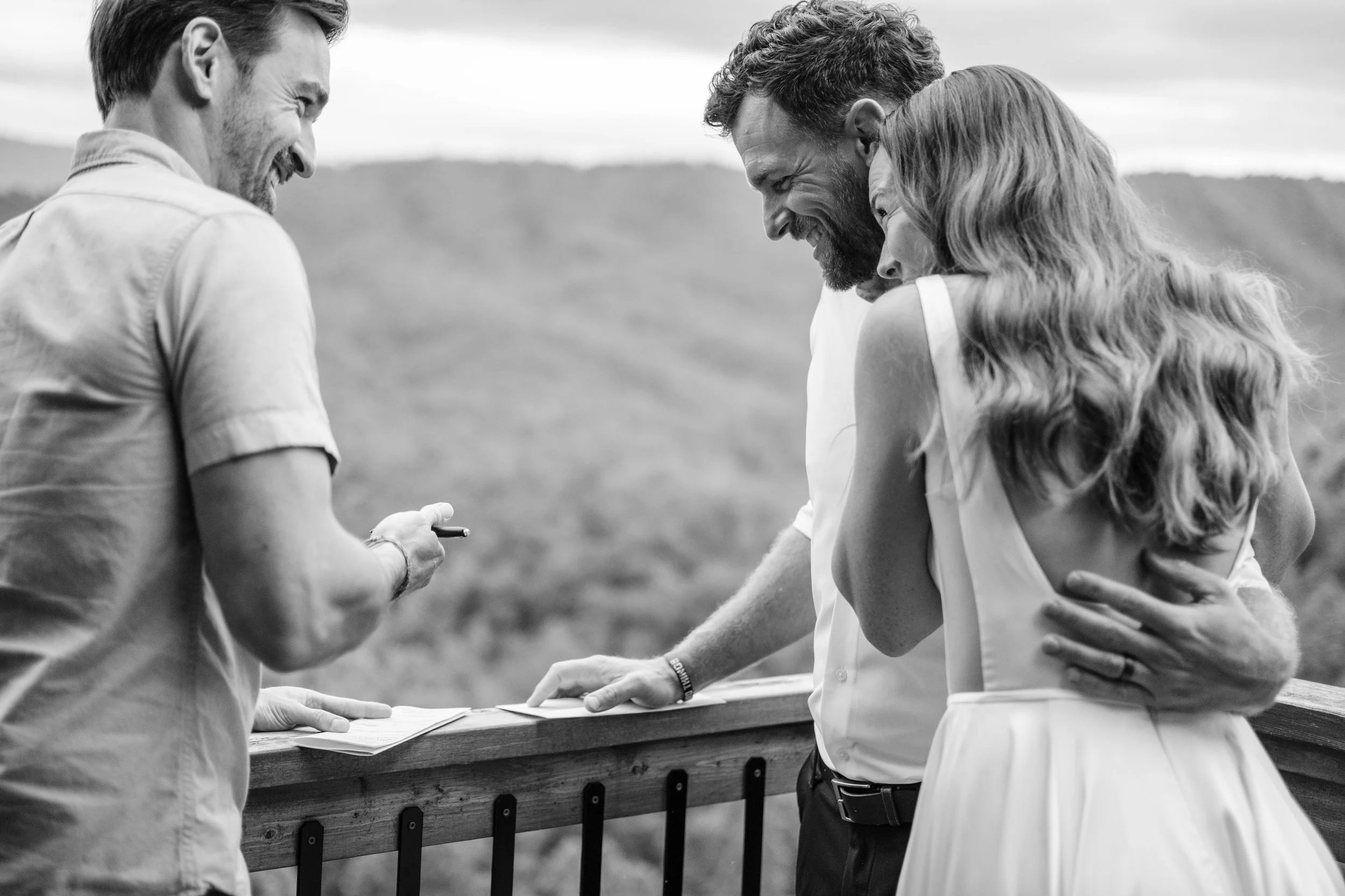A bride and groom embrace as they smile at a man at an outdoor wedding ceremony, with a scenic landscape in the background.
