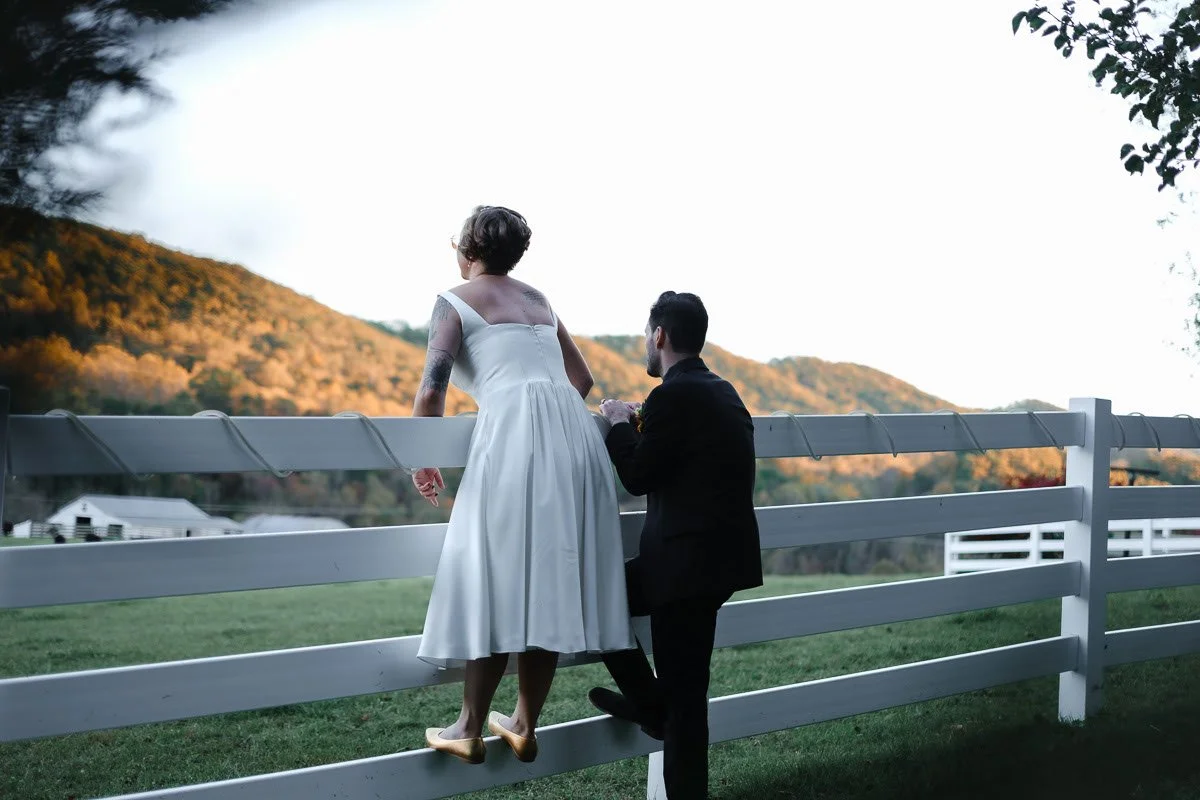 A couple, with the woman in a white dress and the man in a black suit, standing on a white fence in a rural setting, overlooking landscape with hills and trees during sunset.
