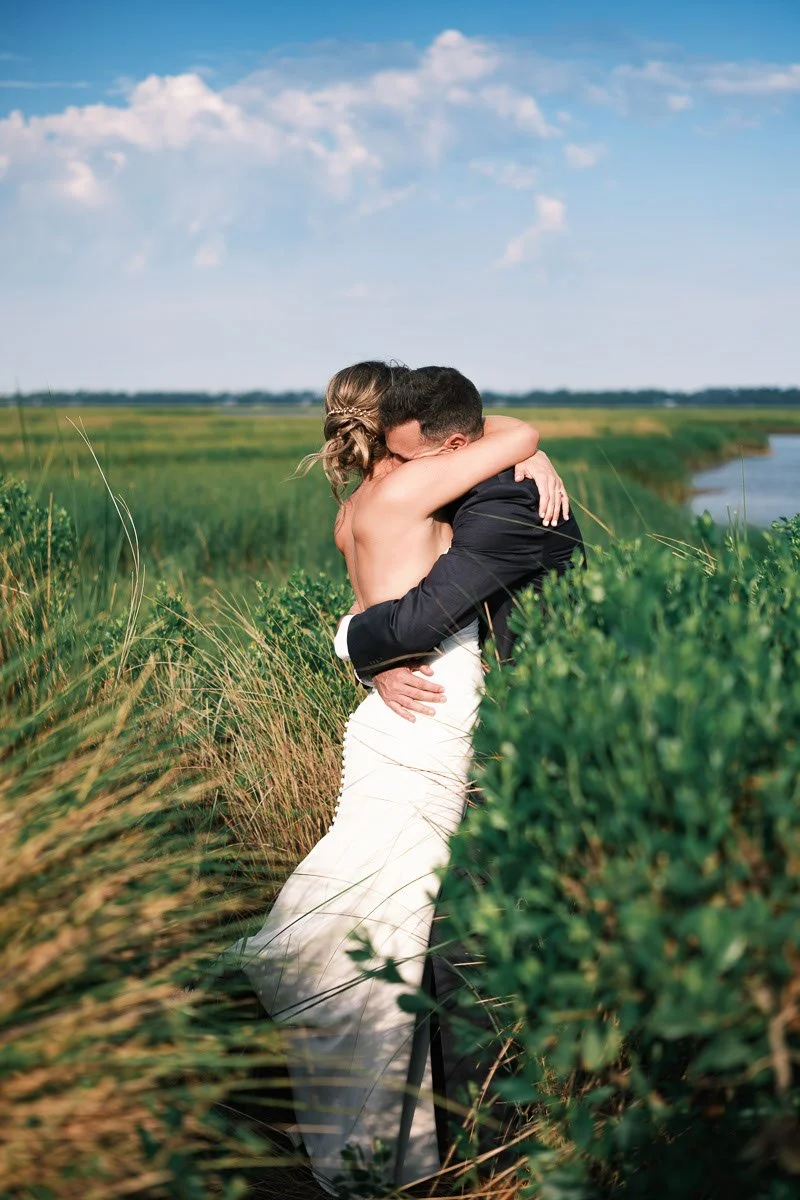 A couple dressed in wedding attire embracing in a lush, green outdoor setting with a river in the background and a partly cloudy sky.