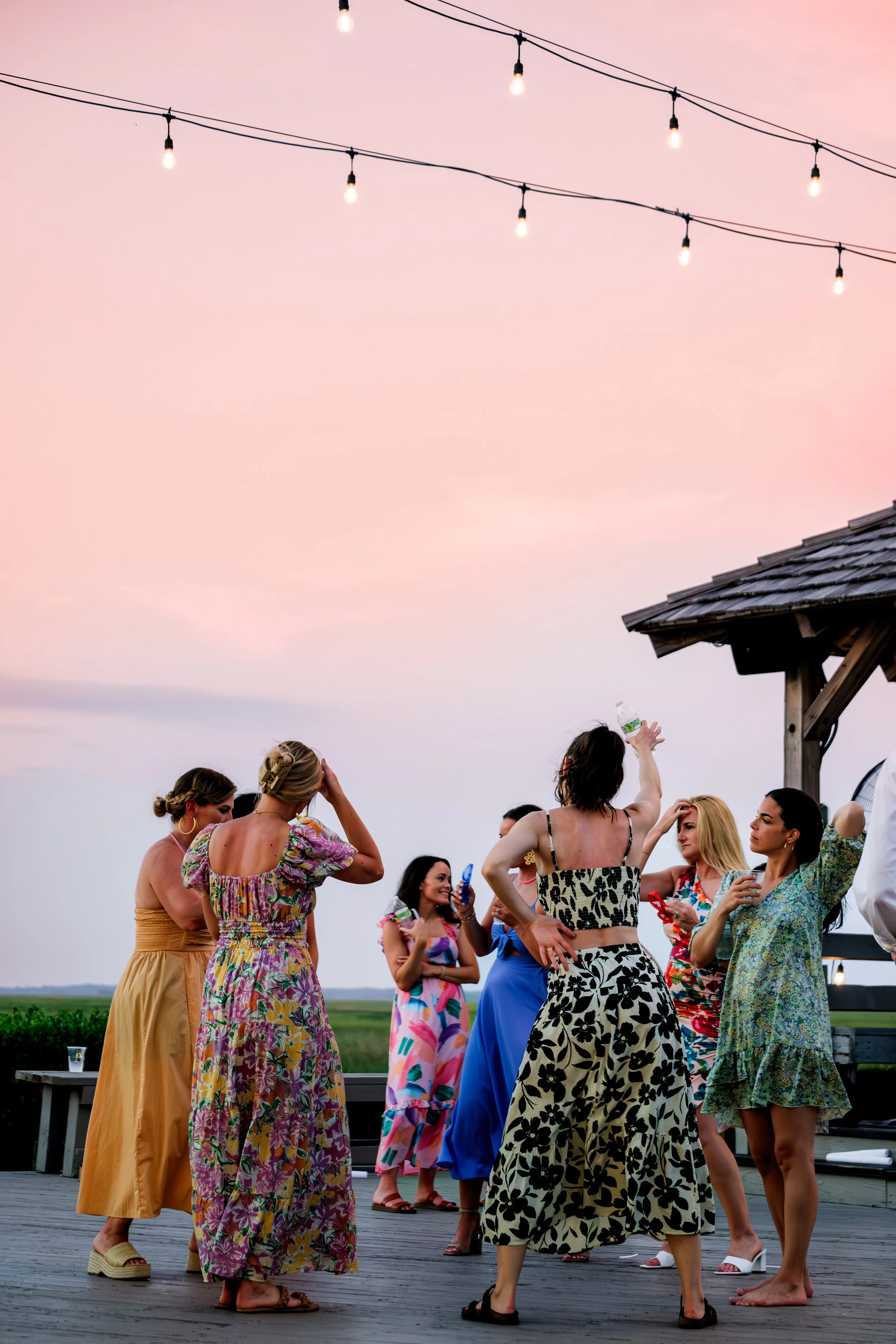 Group of women in colorful dresses gathering outdoors during sunset, under string lights, with a wooden pavilion nearby.