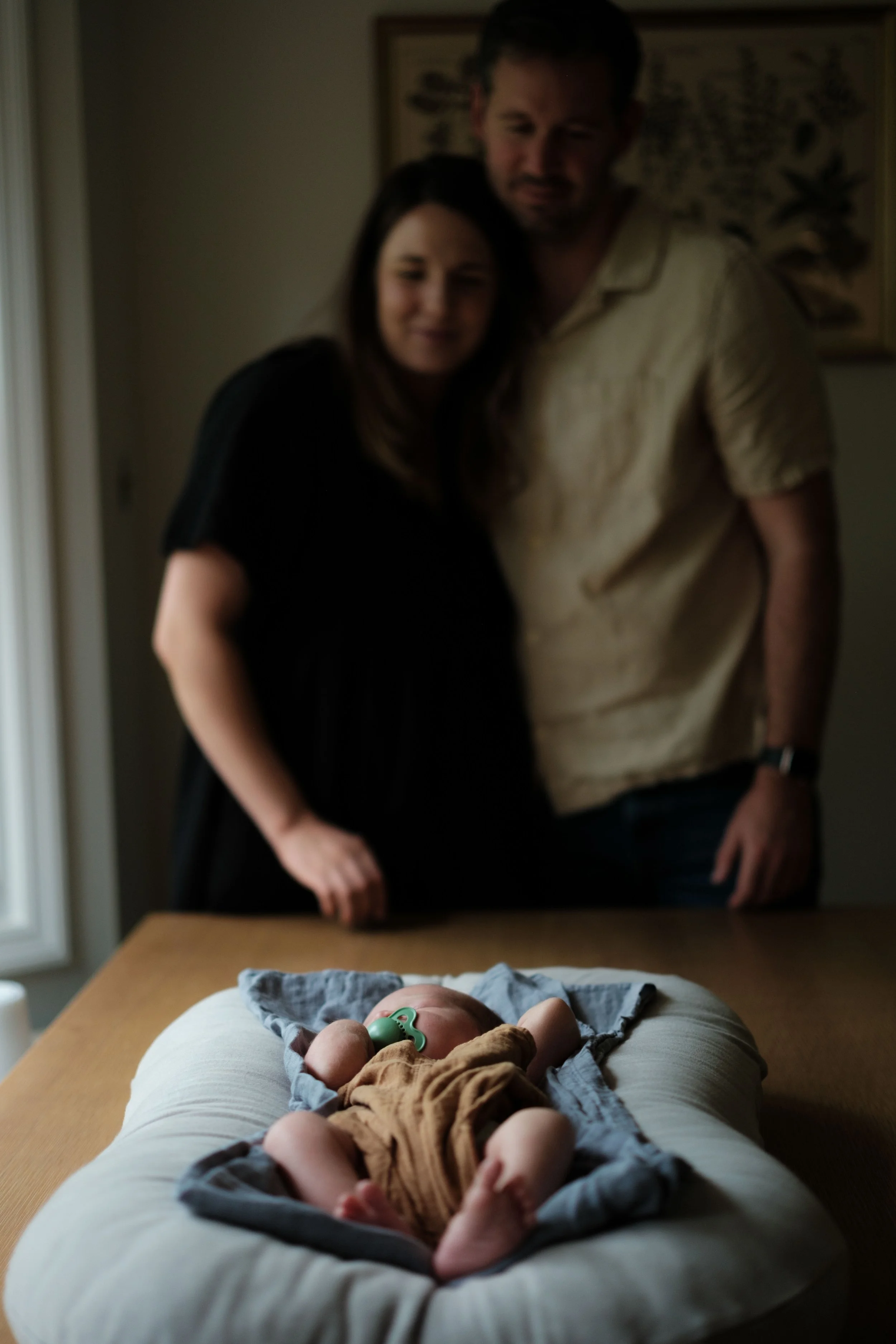 A newborn baby wearing a brown outfit and a green pacifier, lying on a cushioned changing pad on a wooden table, with a blurred couple, likely the parents, in the background.