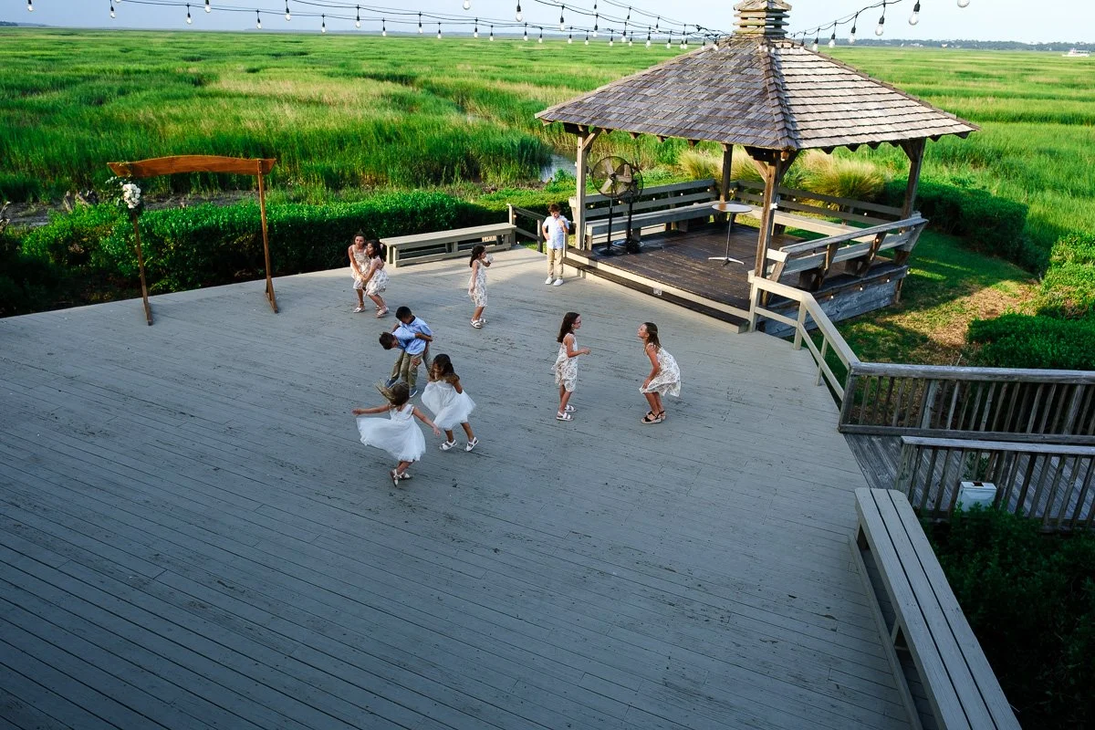 Children dancing and playing on a large wooden deck next to a gazebo with string lights overhead, surrounded by green marshland.