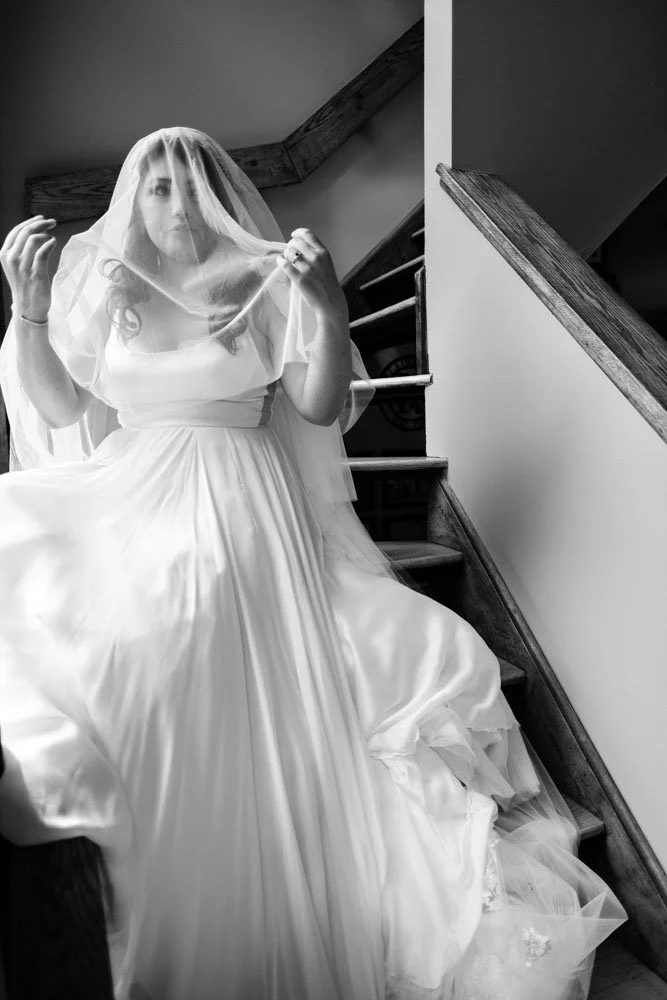 A woman in a wedding dress sitting on stairs and holding a veil over her face.