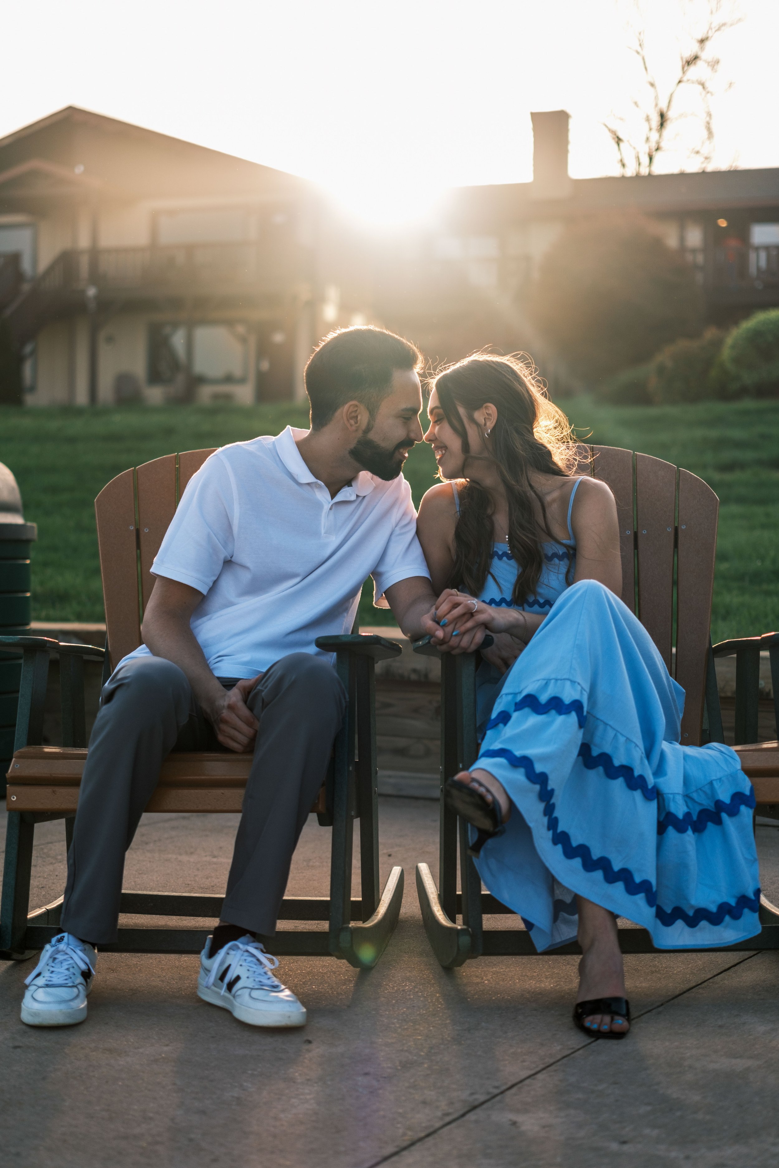 A couple sitting on wooden outdoor chairs holding hands, leaning their foreheads together at sunset in a backyard, with a house and trees in the background.
