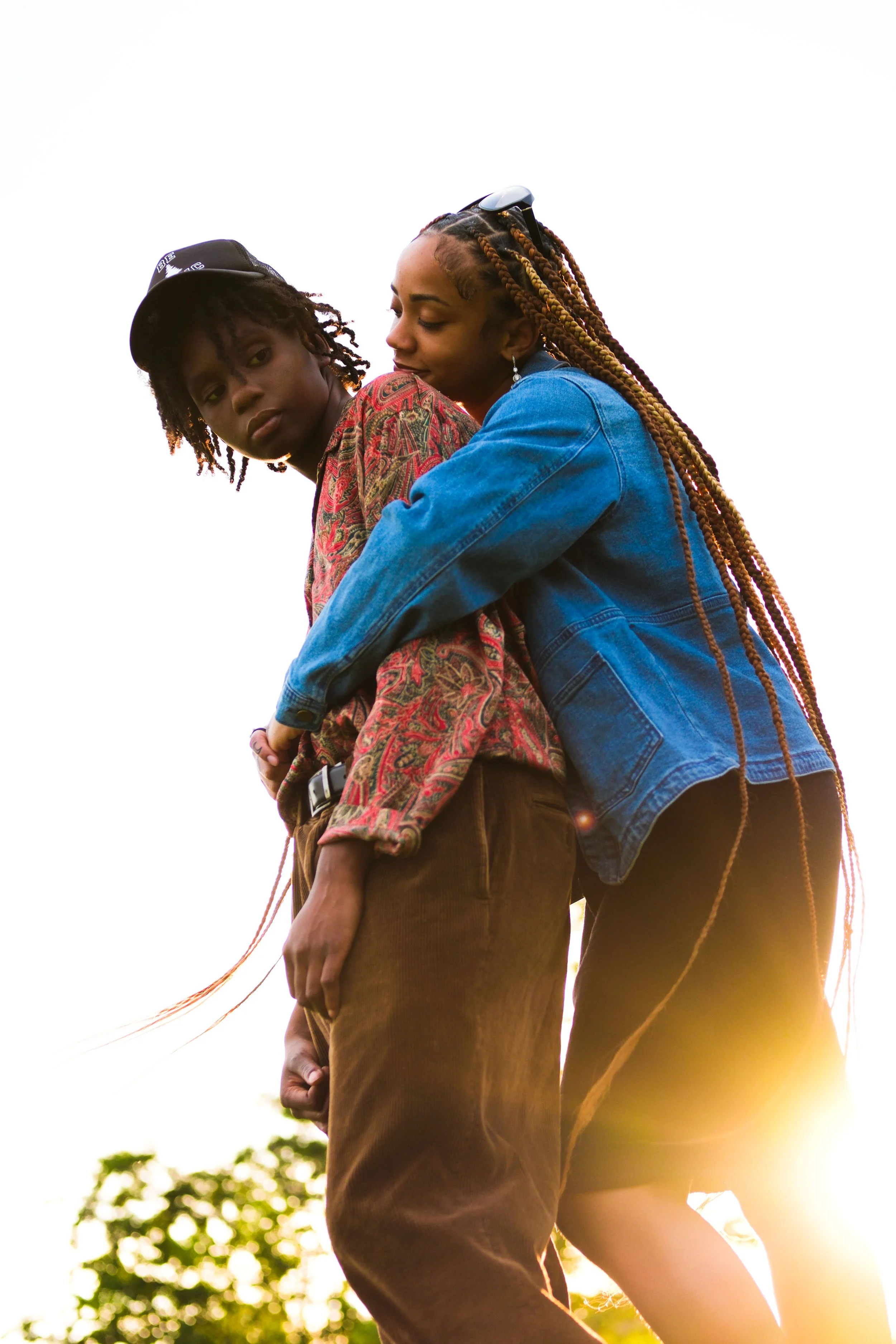 A woman with long braided hair and sunglasses on her head hugging a young person in a colorful jacket from behind outdoors at sunset.