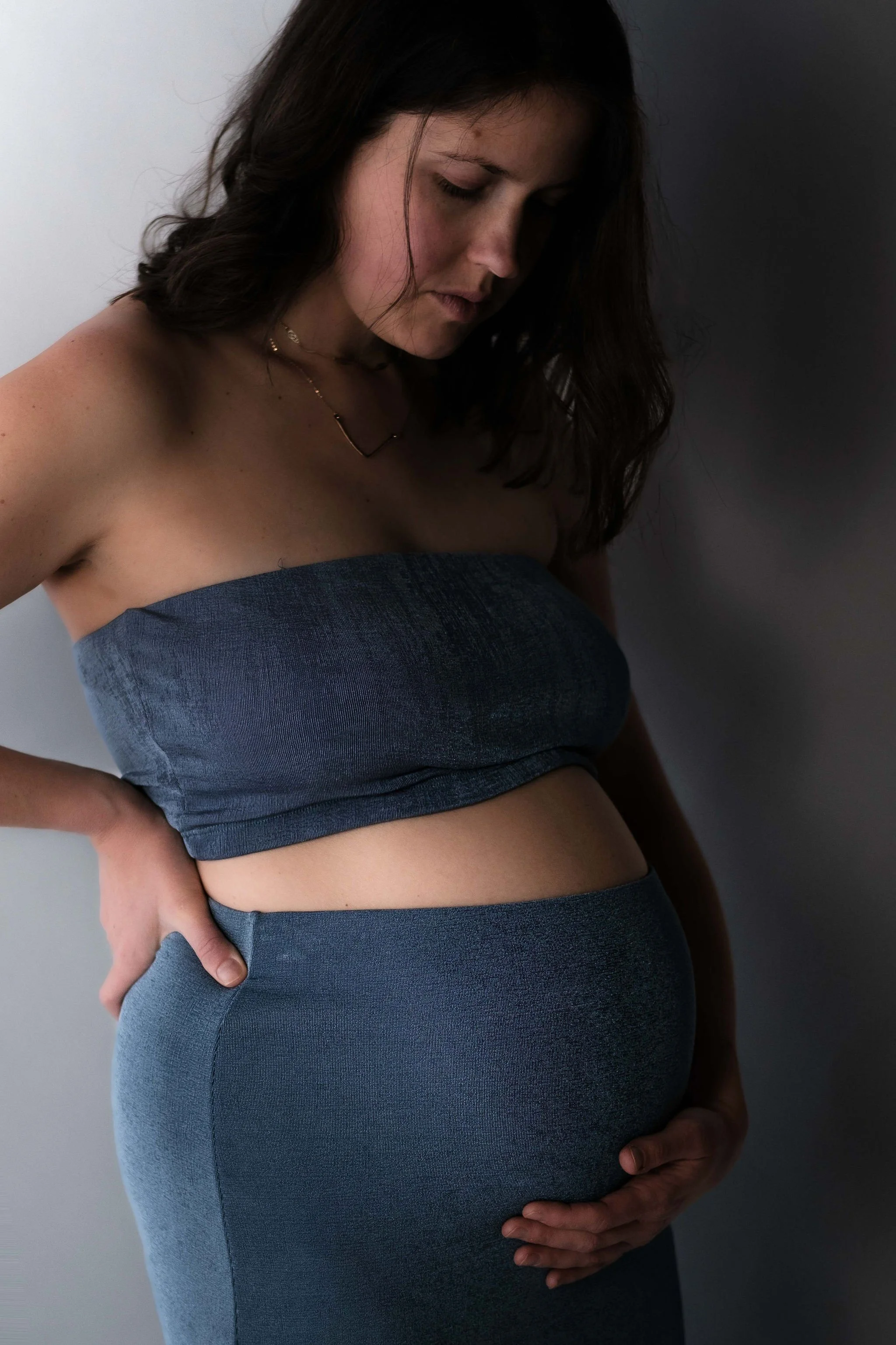 A pregnant woman with shoulder-length dark hair, wearing a strapless blue top and matching blue leggings, standing against a faded gray background, gently holding her belly.