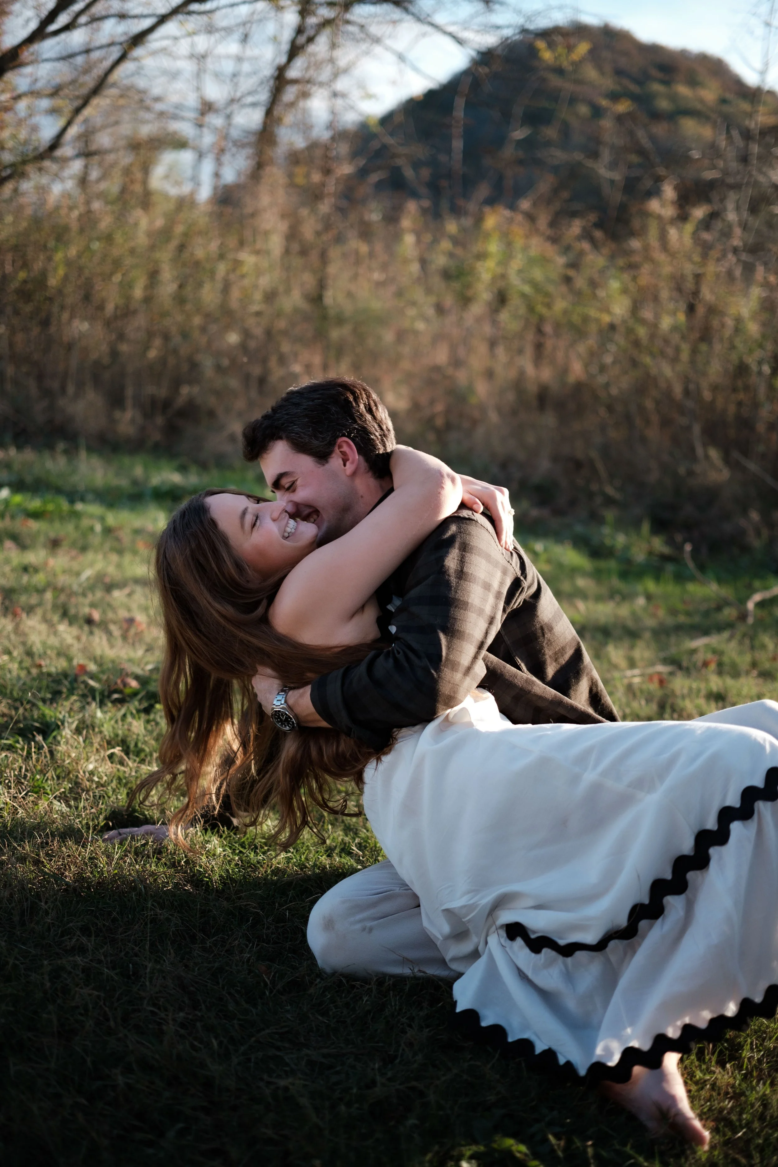 A couple hugging and smiling outdoors on grass with trees and a mountain in the background during daylight