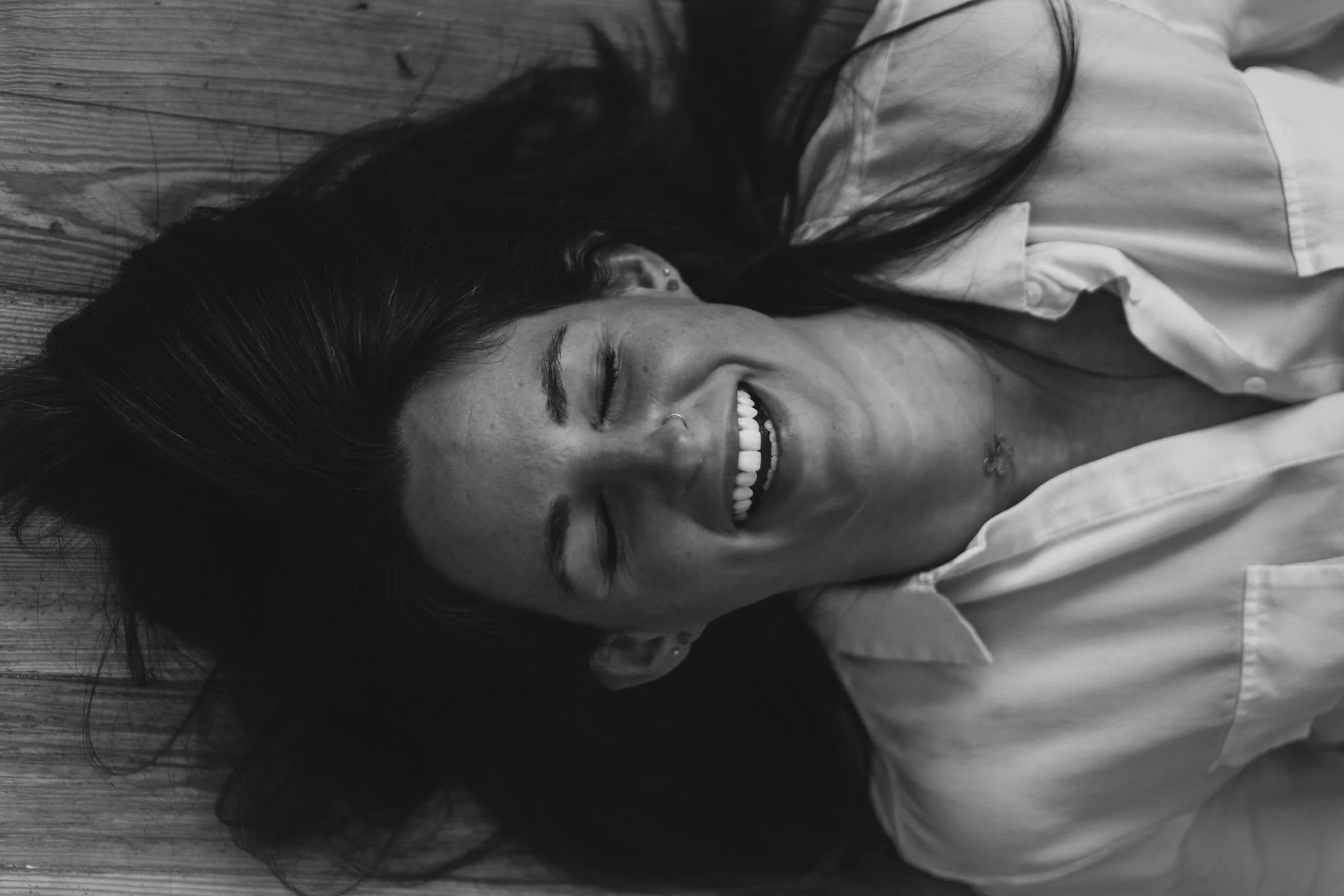 A woman with long dark hair lying on a wooden floor, smiling with her eyes closed, wearing a white shirt.