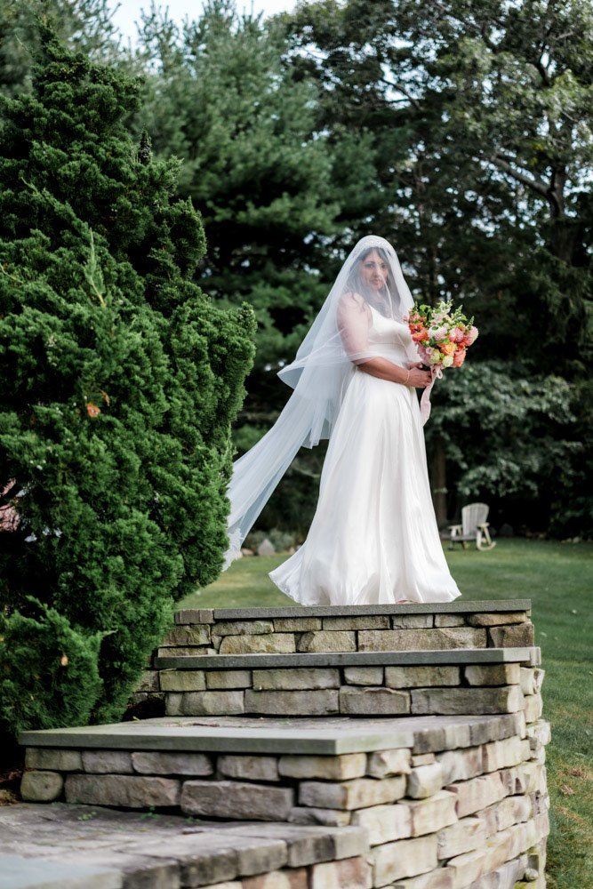 Bride in a white wedding dress with a veil holding a bouquet of pink and white flowers standing on a stone staircase outdoors surrounded by greenery.
