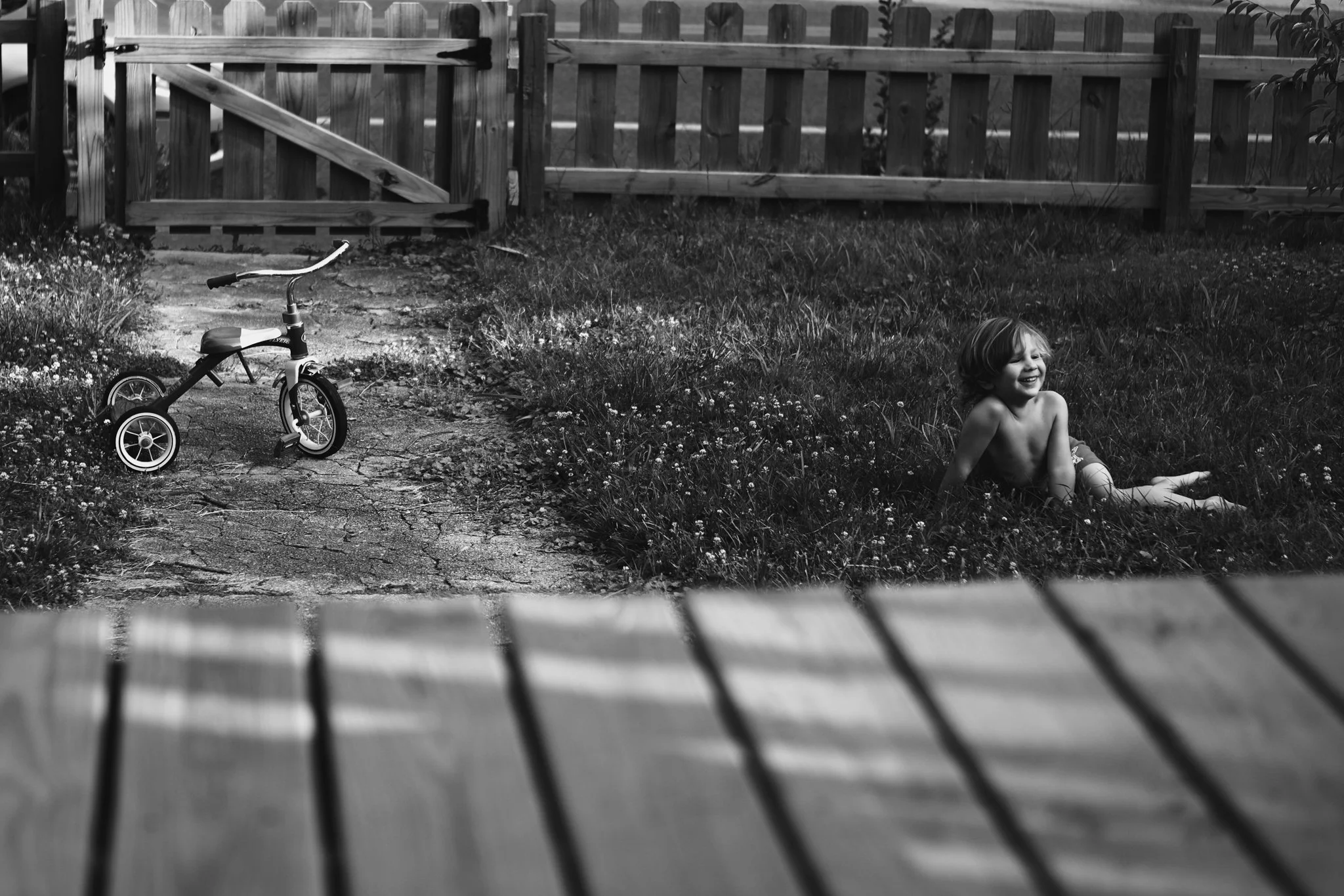A young child sitting in the grass and smiling, with a small bicycle with training wheels on a cracked path nearby, enclosed by a wooden fence.