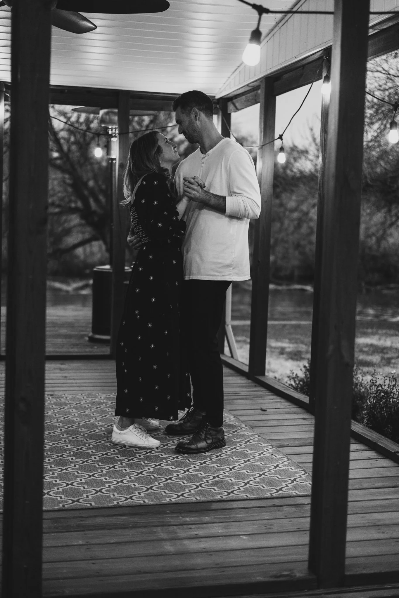 A couple dancing closely on a porch decorated with string lights, with trees in the background.