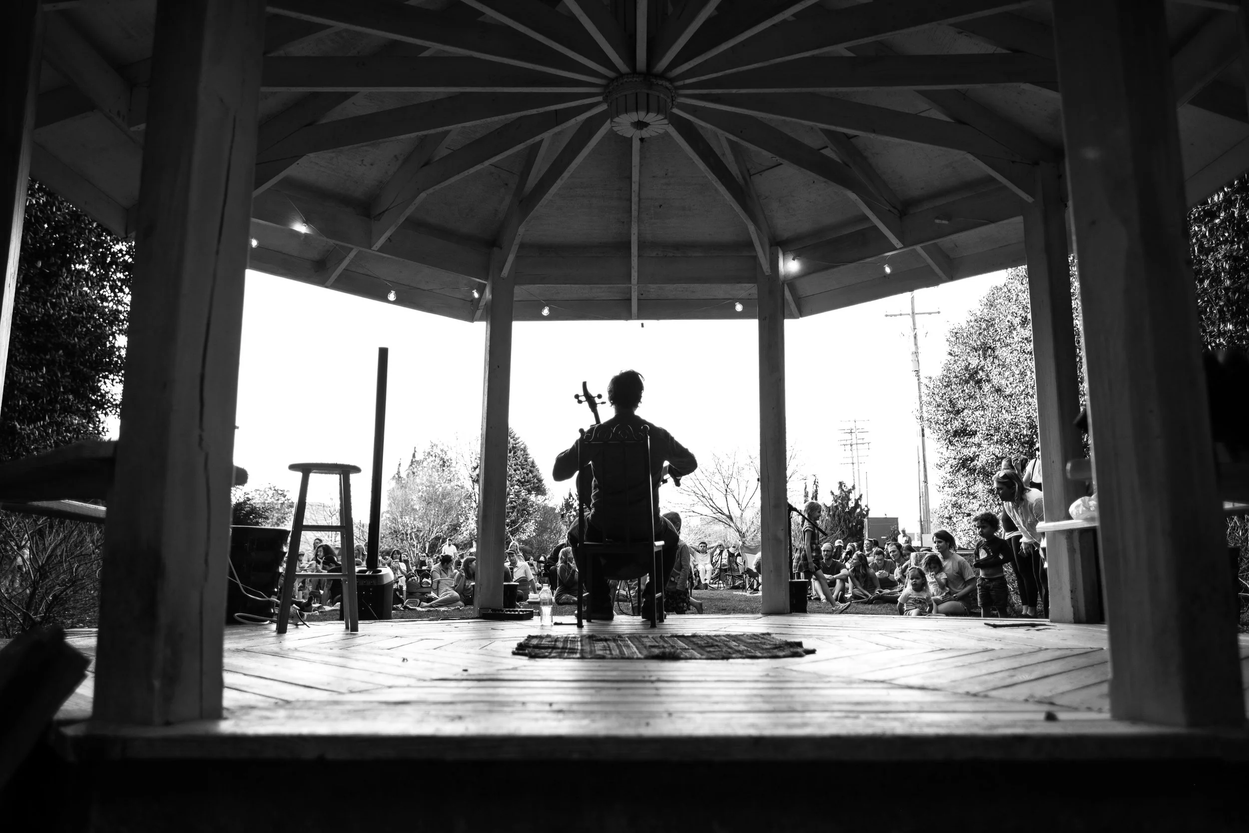 A person playing a musical instrument on a stage in front of an audience outdoors, viewed from beneath a gazebo structure.