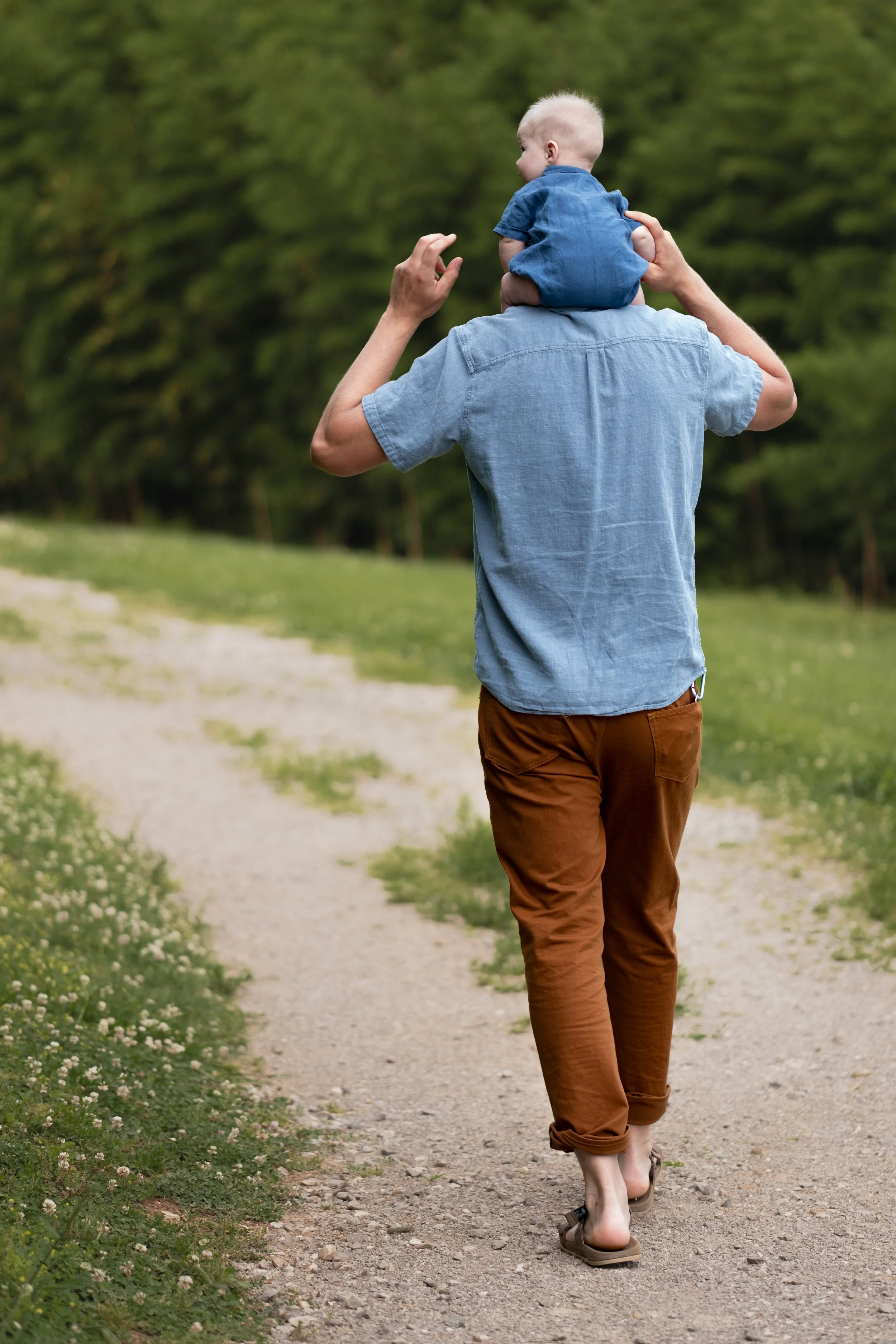 A person carrying a young child on their shoulders while walking on a dirt path surrounded by greenery.