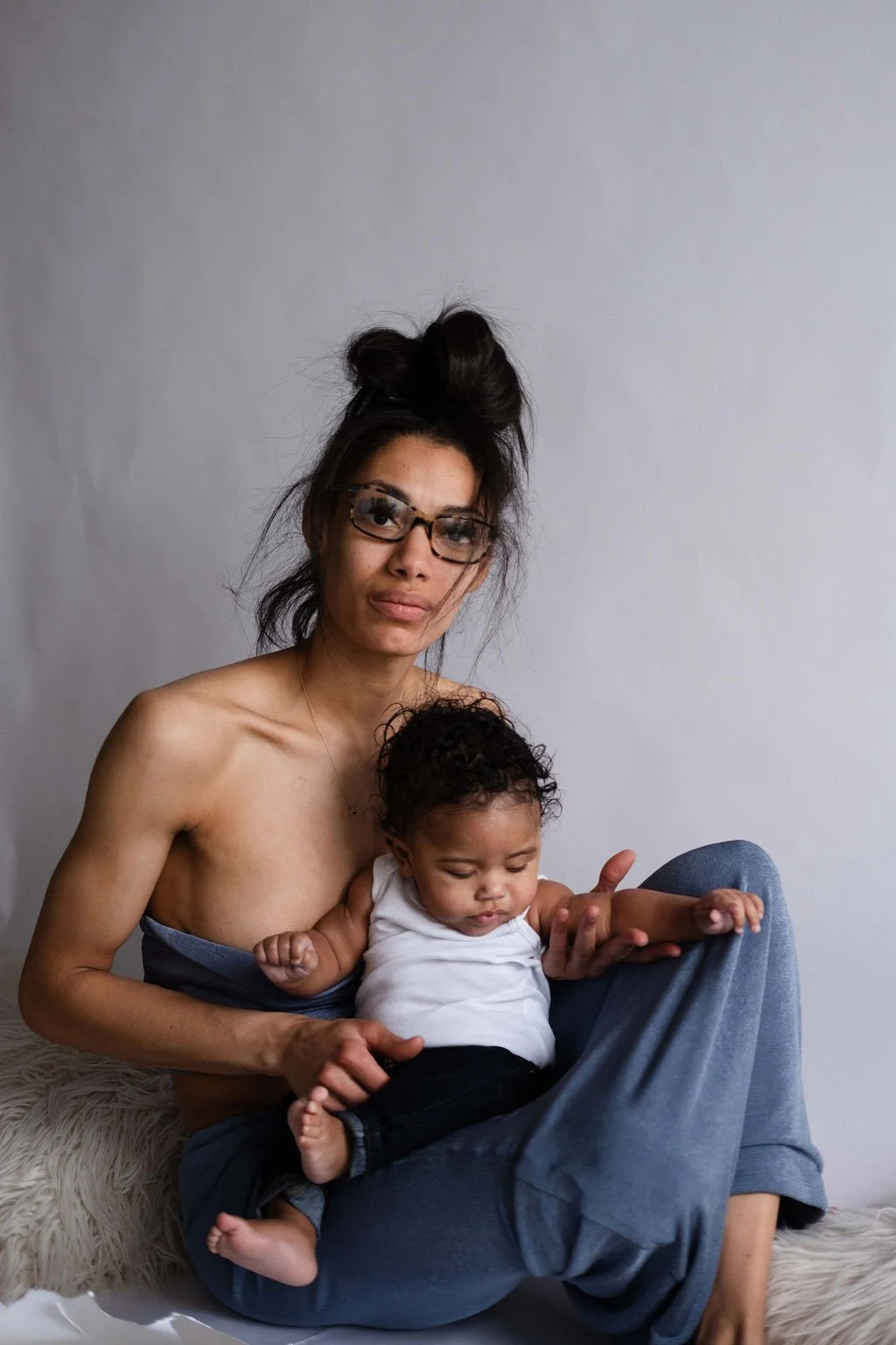 A woman with glasses and a messy bun holding a baby sitting on her lap, both sitting on a soft white surface against a plain light gray background.
