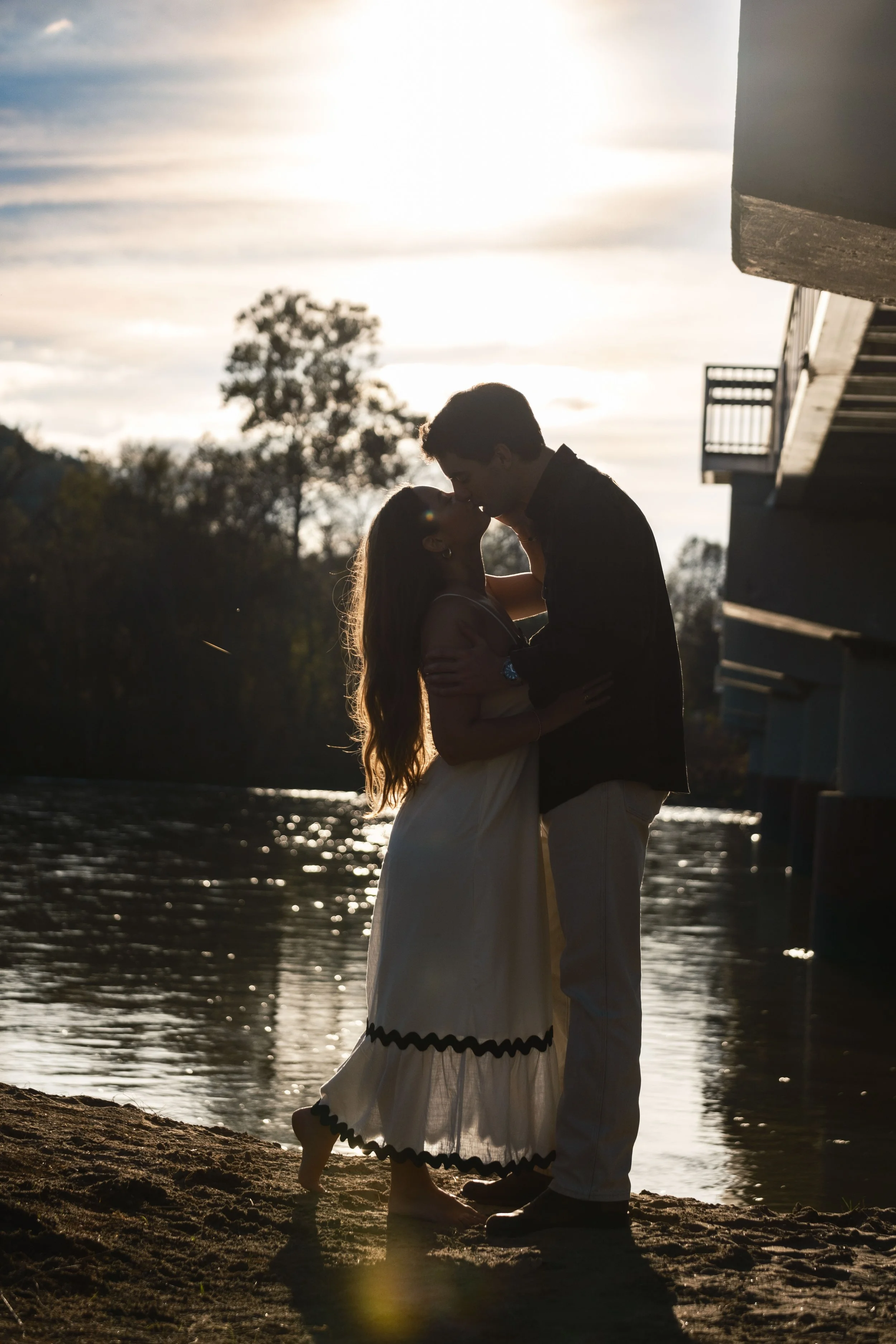 Silhouette of a couple kissing near the water at sunset with trees and a building in the background.
