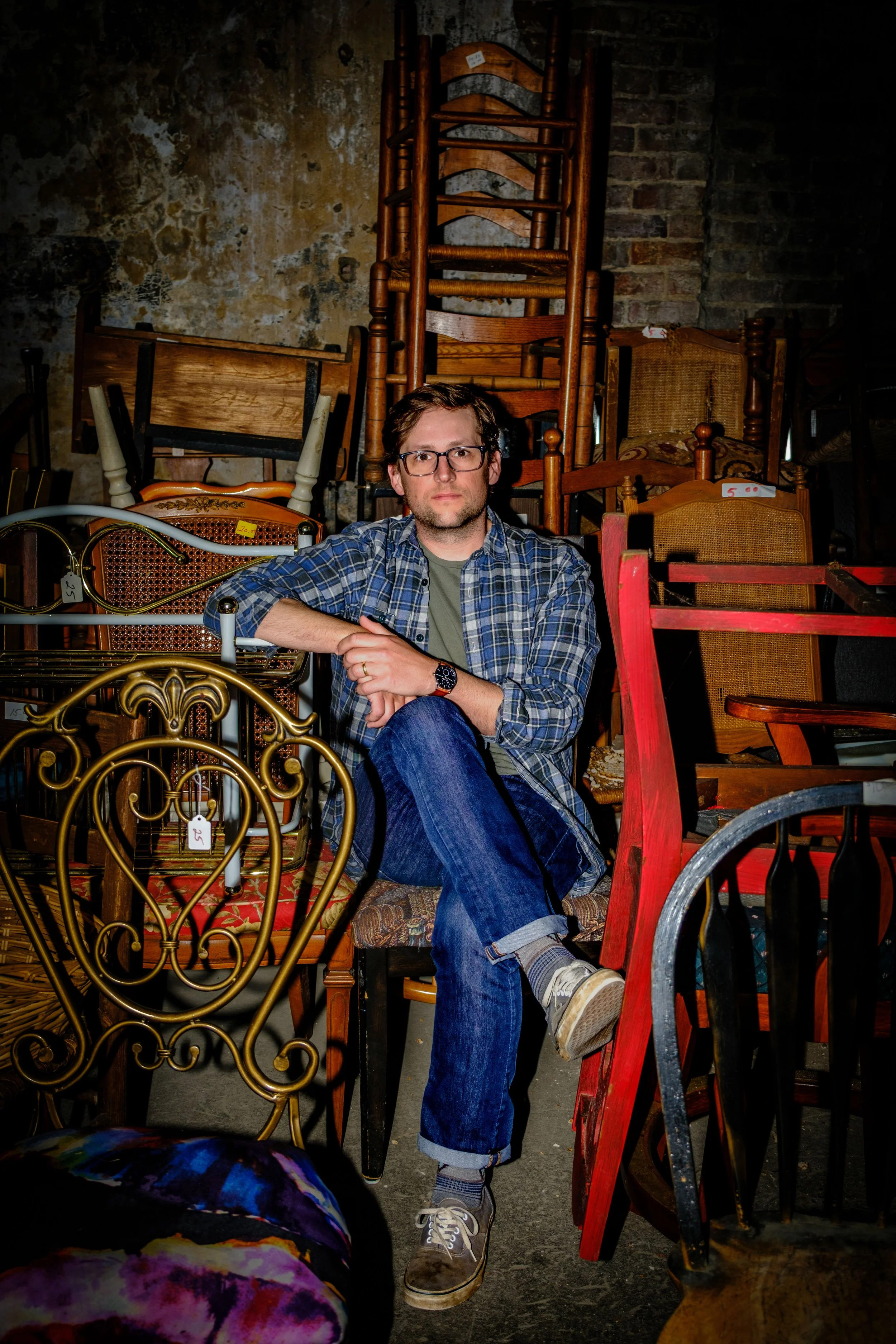 A man with glasses and a plaid shirt sitting amidst a collection of various chairs and furniture in a cluttered space.