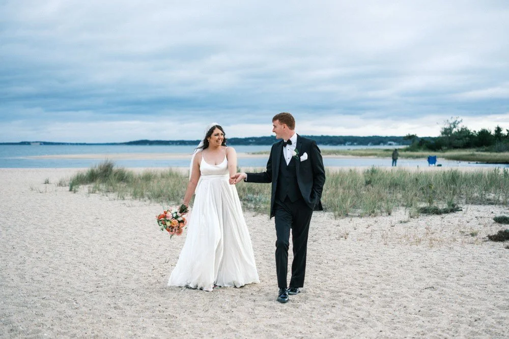 Bride and groom walking hand in hand on a beach, with the bride holding a bouquet of flowers, the groom in a black tuxedo, and the background showing water, sand, and some people in the distance.