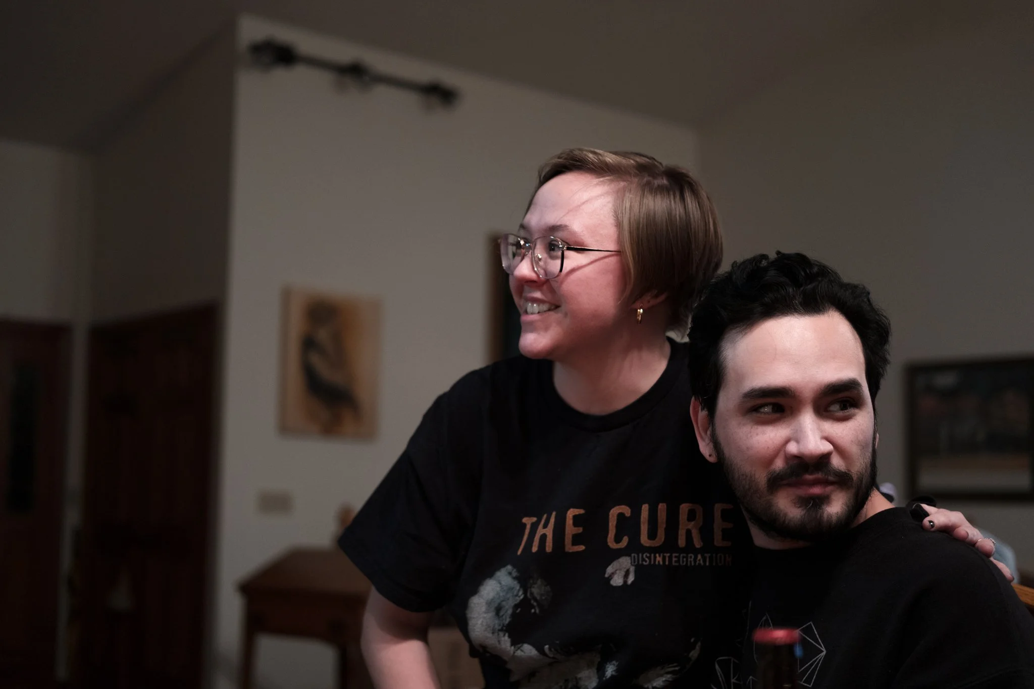 Two people sitting close together indoors, one with short blonde hair wearing glasses and a black The Cure t-shirt, the other with dark hair and a beard, wearing a black shirt. They are smiling and appear to be happy.