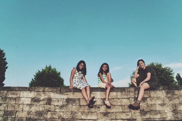 Three girls sitting on top of a stone wall outdoors, with trees and a blue sky in the background.
