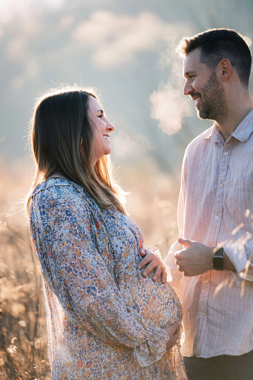 A pregnant woman and a man stand face to face outdoors, smiling at each other during sunset. The woman is wearing a floral dress and has her hands on her belly, while the man is dressed in a light-colored, striped button-up shirt and wears a smartwat