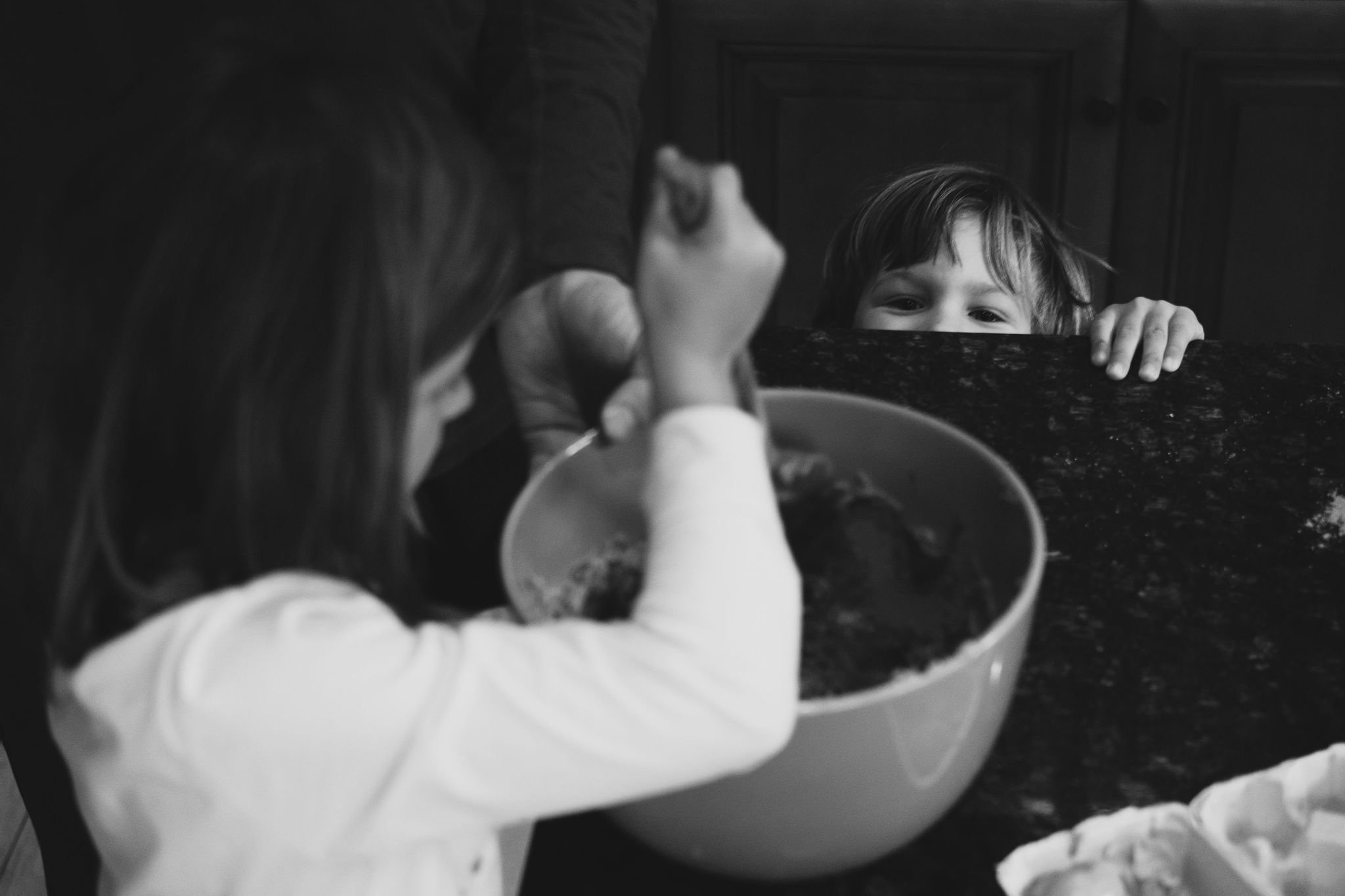 A child with glasses eating from a large bowl at a dining table, with another child peeking over the edge of the table, looking at the first child with a smile.