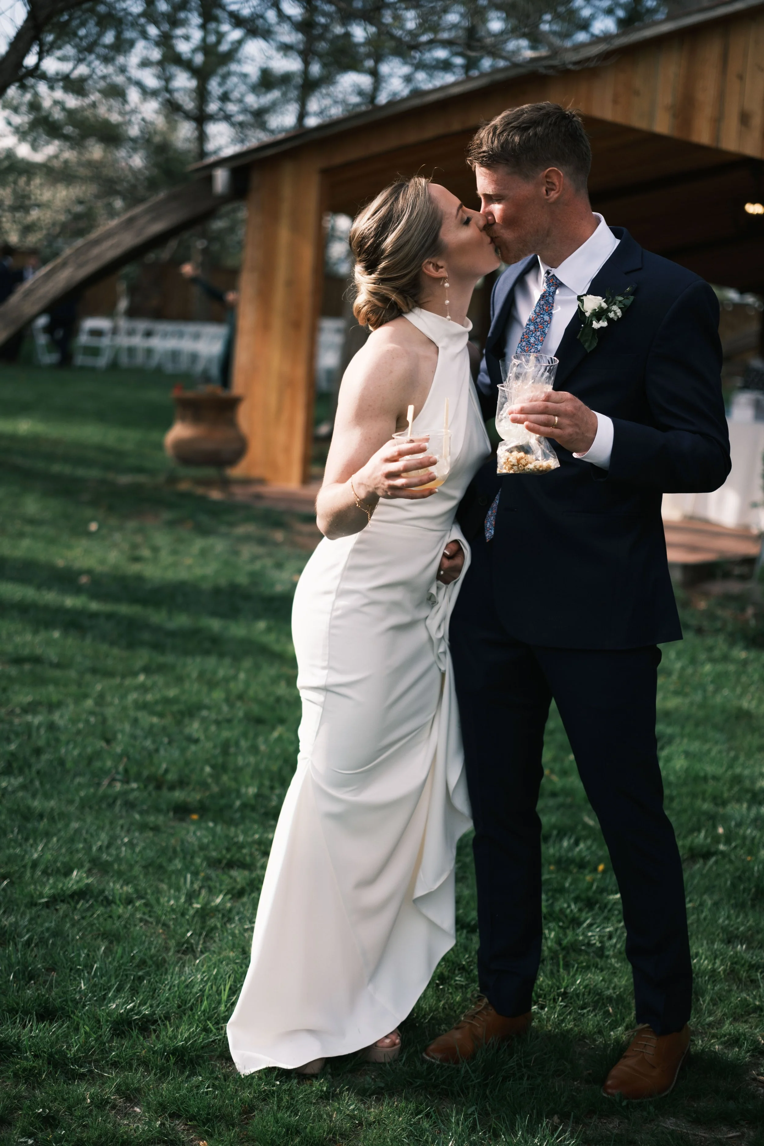 A bride and groom sharing a kiss at their outdoor wedding reception, standing on green grass with a wooden building in the background.