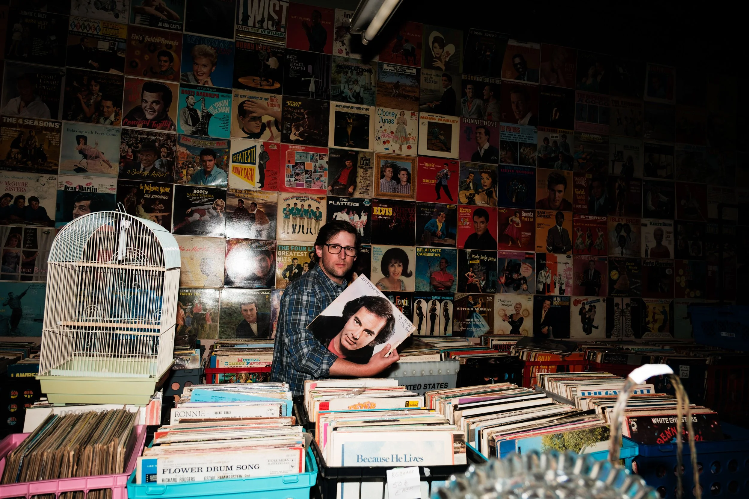 Man shopping for records at a vinyl store, holding a record with Elvis Presley on the cover, surrounded by shelves of vinyl records and a wall decorated with vintage album covers.