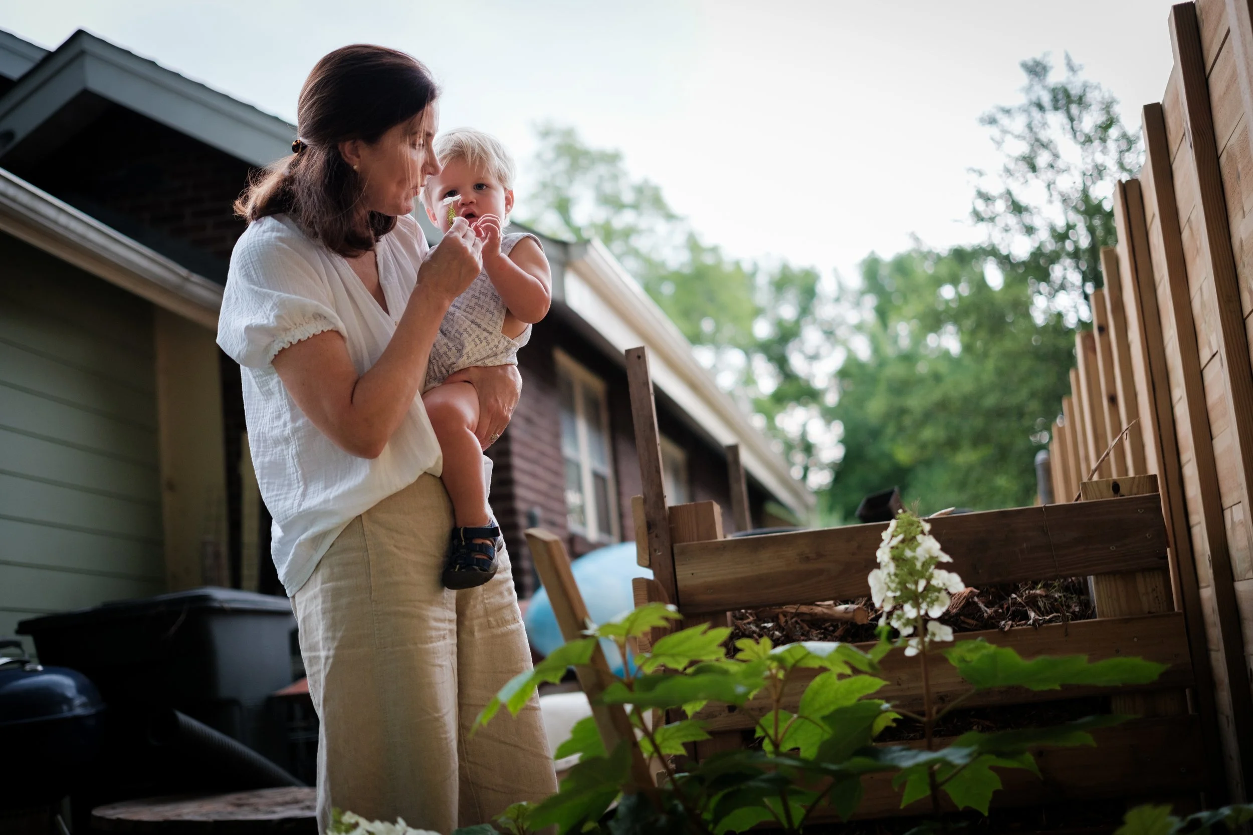 A woman holding a young child in a backyard, both looking at a flower in a wooden garden bed.