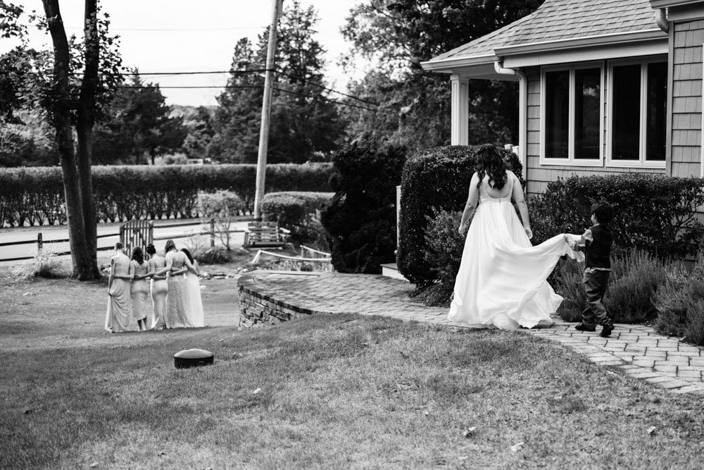 A woman in a wedding dress walking on a brick pathway holding hands with a young boy, with three women in dresses standing in the background near a house.