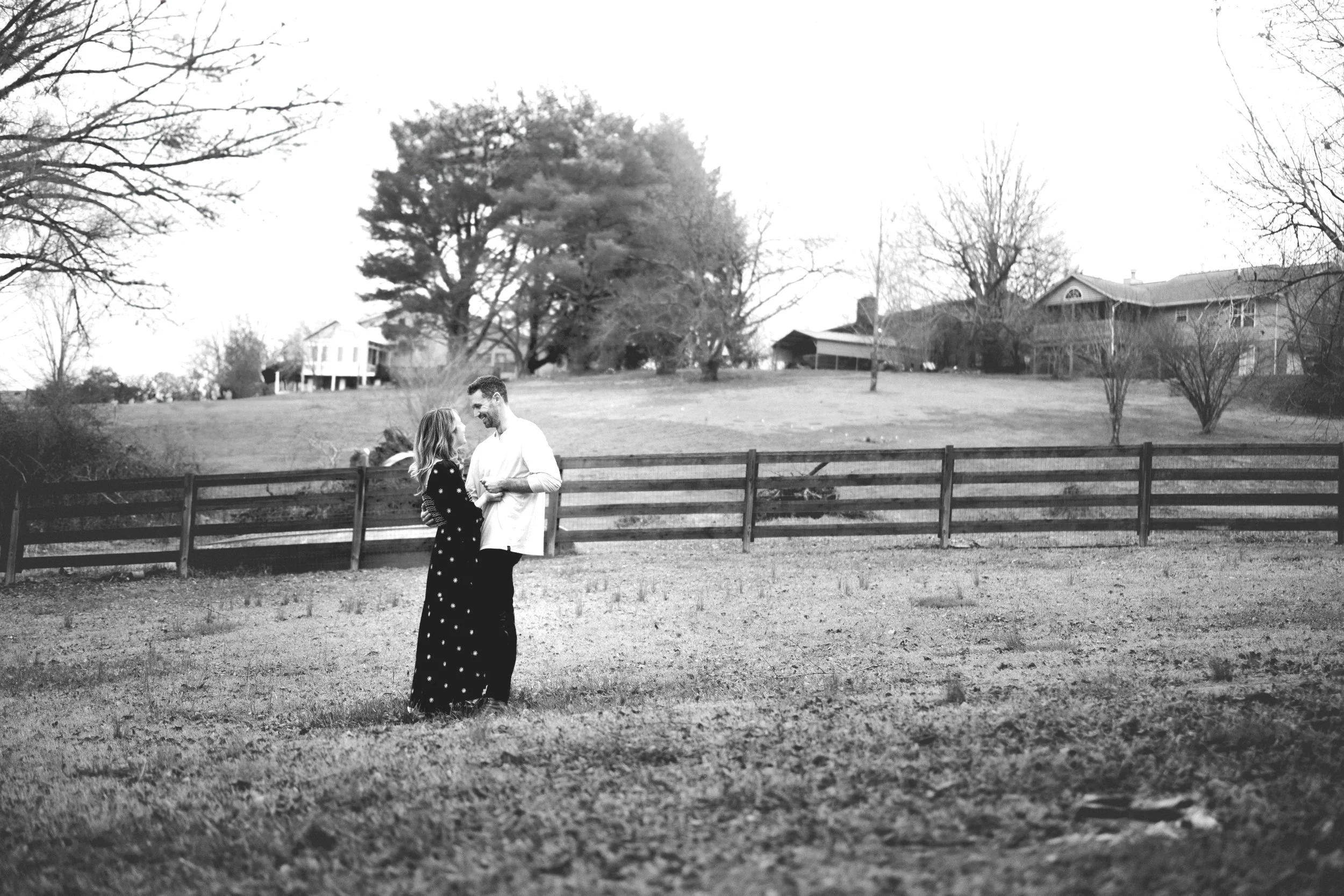 A man and woman standing in a grassy yard, facing each other and smiling, with a wooden fence and houses and trees in the background.