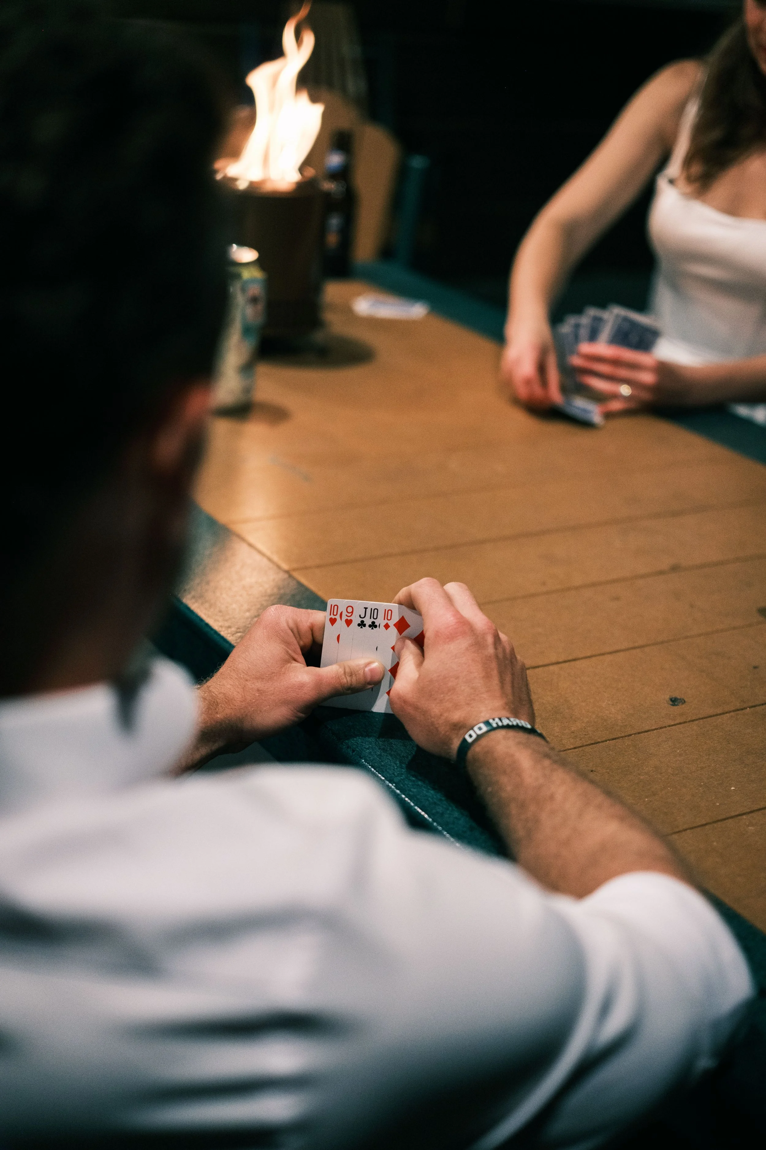 People playing poker at a wooden table, with a man holding a hand of five cards and a woman in the background holding poker chips.
