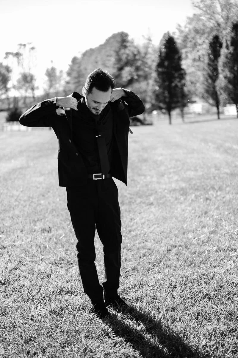 A man in a suit adjusting his jacket outdoors on grass, with trees in the background.