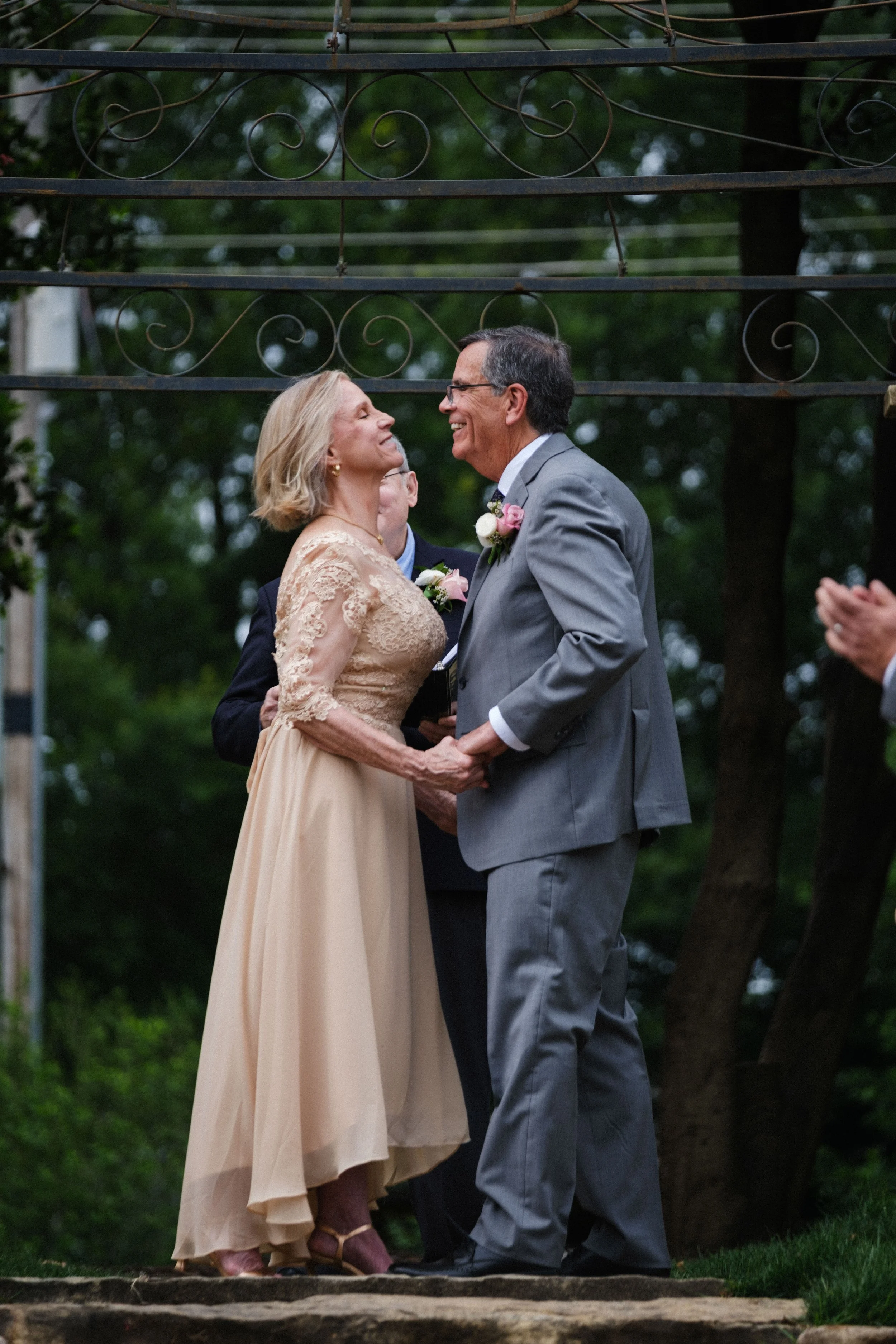 An elderly couple holding hands and smiling at each other, outdoors during a wedding ceremony, with a person clapping to the right and greenery in the background.