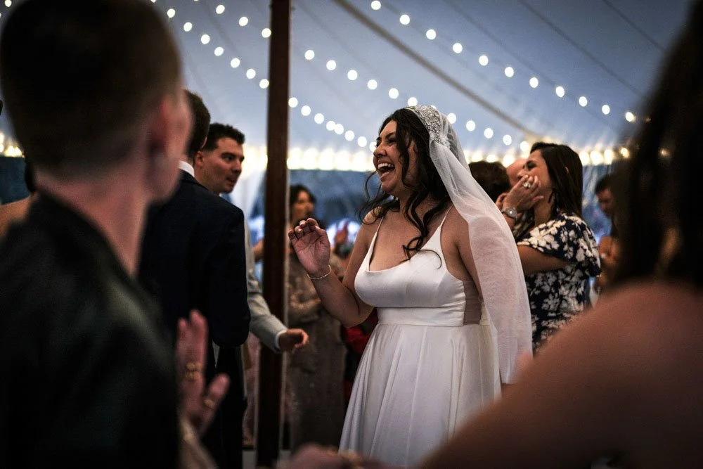 Bride in a white wedding dress and veil laughing with friends at a celebration under a decorated tent.