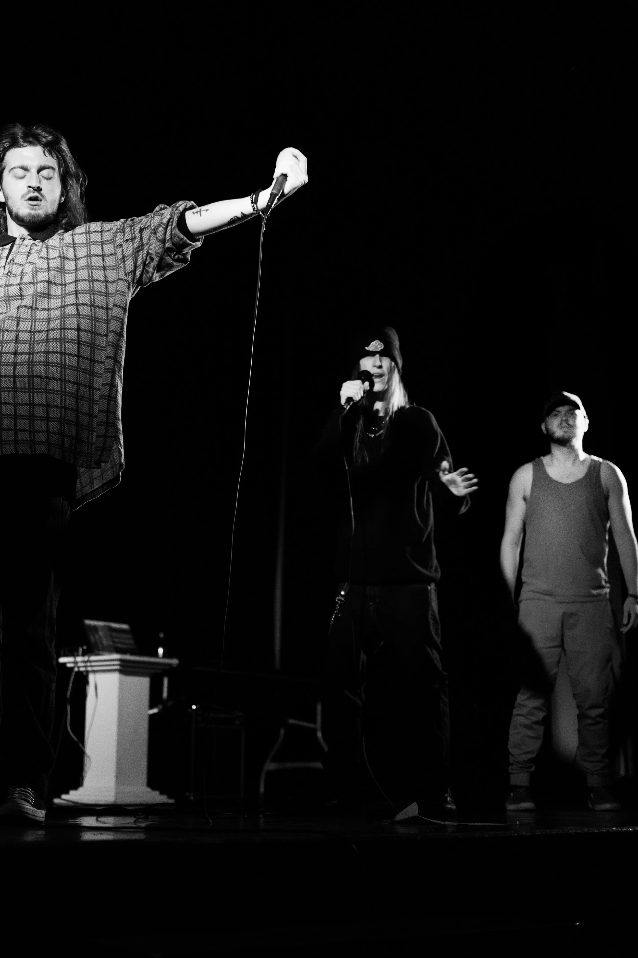 Three people performing on a dark stage with microphones, all casually dressed, in black and white.