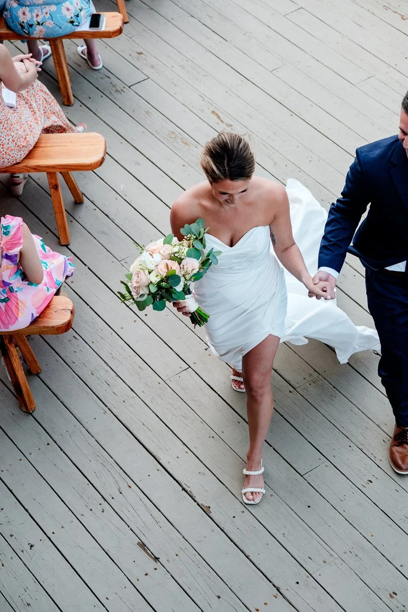 Wedding bride in white dress holding a bouquet, walking on wooden deck, holding hands with a man in a blue suit.