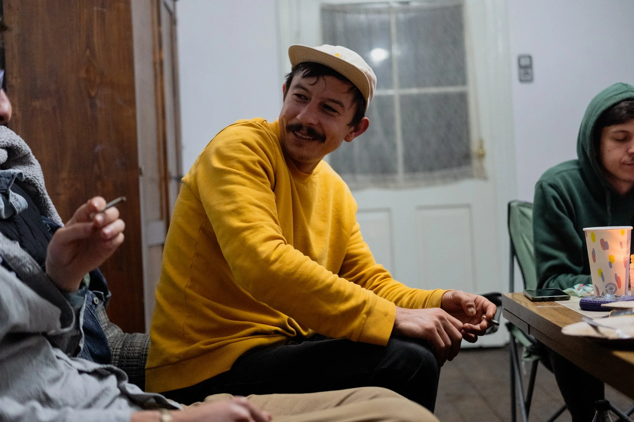 A group of young men sitting at a table, engaged in conversation. The man in the center wearing a yellow sweatshirt and a beige cap is smiling and looking to his left. The table has a few items, including a colorful cup and a phone. The background sh