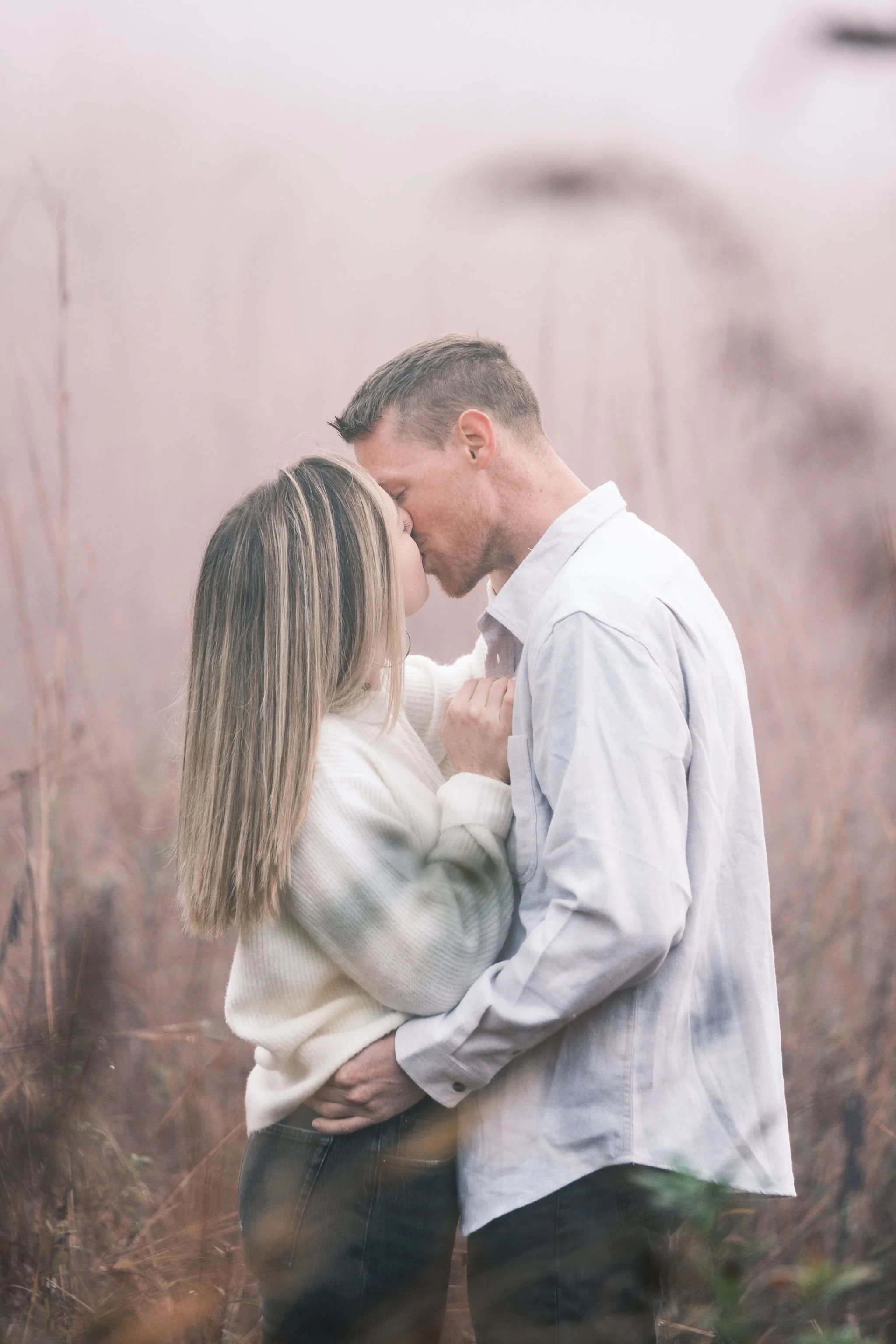 A couple sharing a romantic kiss outdoors in a field of tall, brown grasses with a soft, pinkish sky.