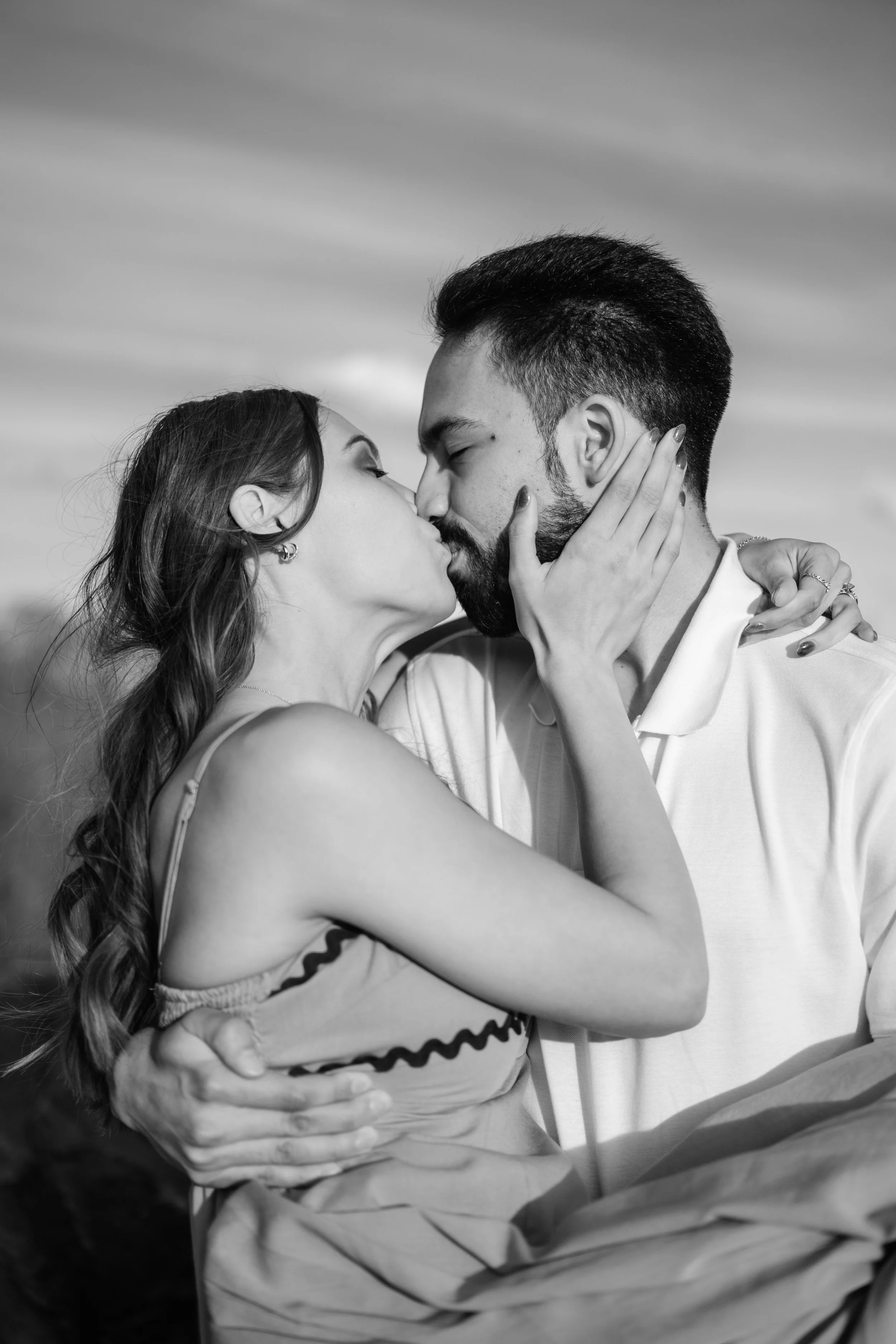 A black and white photo of a couple kissing outdoors, with the woman holding the man's face and the man embracing her.