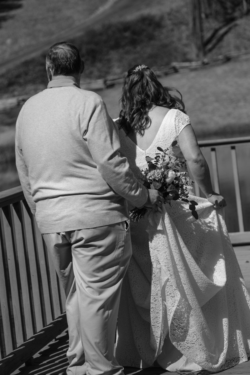 A bride holding a bouquet of flowers and holding up her wedding dress, standing next to a man on a wooden deck outdoors.