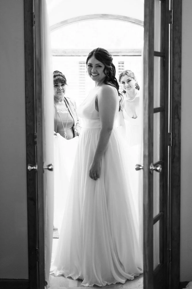 A woman in a wedding dress standing in a doorway with two women behind her, all smiling in a bright room.